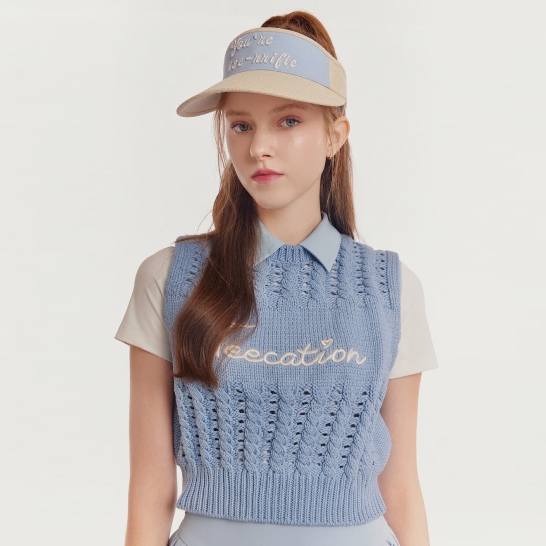 A woman wearing a light blue knit polo vest over a cream short-sleeve shirt, paired with a beige visor, against a plain light gray studio background.