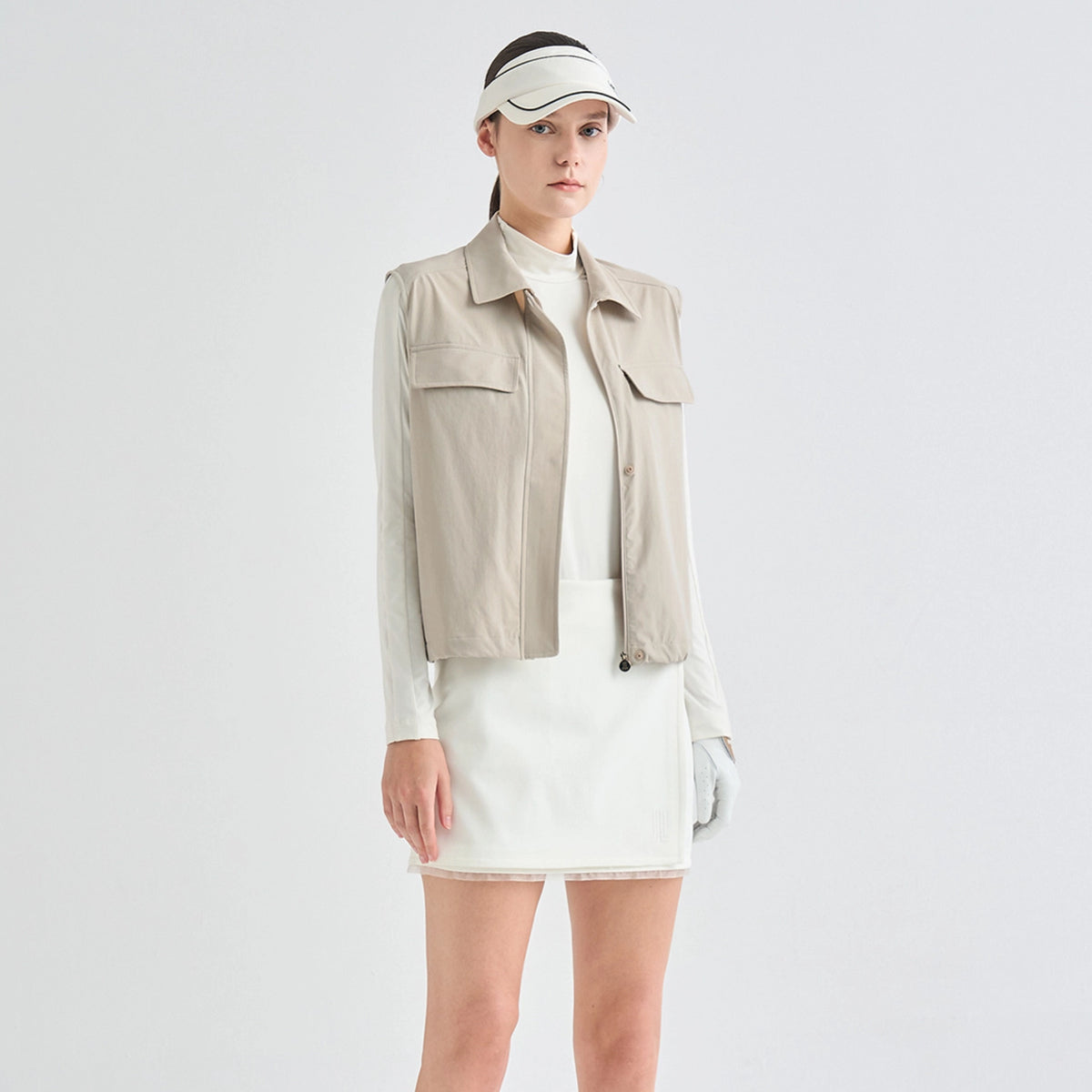 A model wearing a beige sleeveless utility jacket over a white long-sleeve top and white mini skirt, paired with a white visor and white gloves, against a light gray studio background.