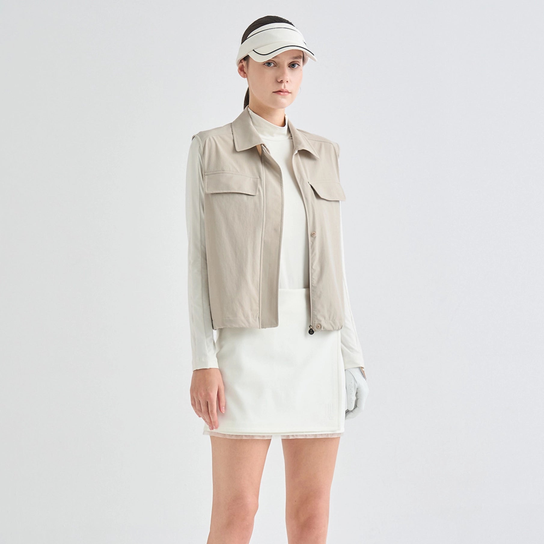 A model wearing a beige sleeveless utility jacket over a white long-sleeve top and white mini skirt, paired with a white visor and white gloves, against a light gray studio background.