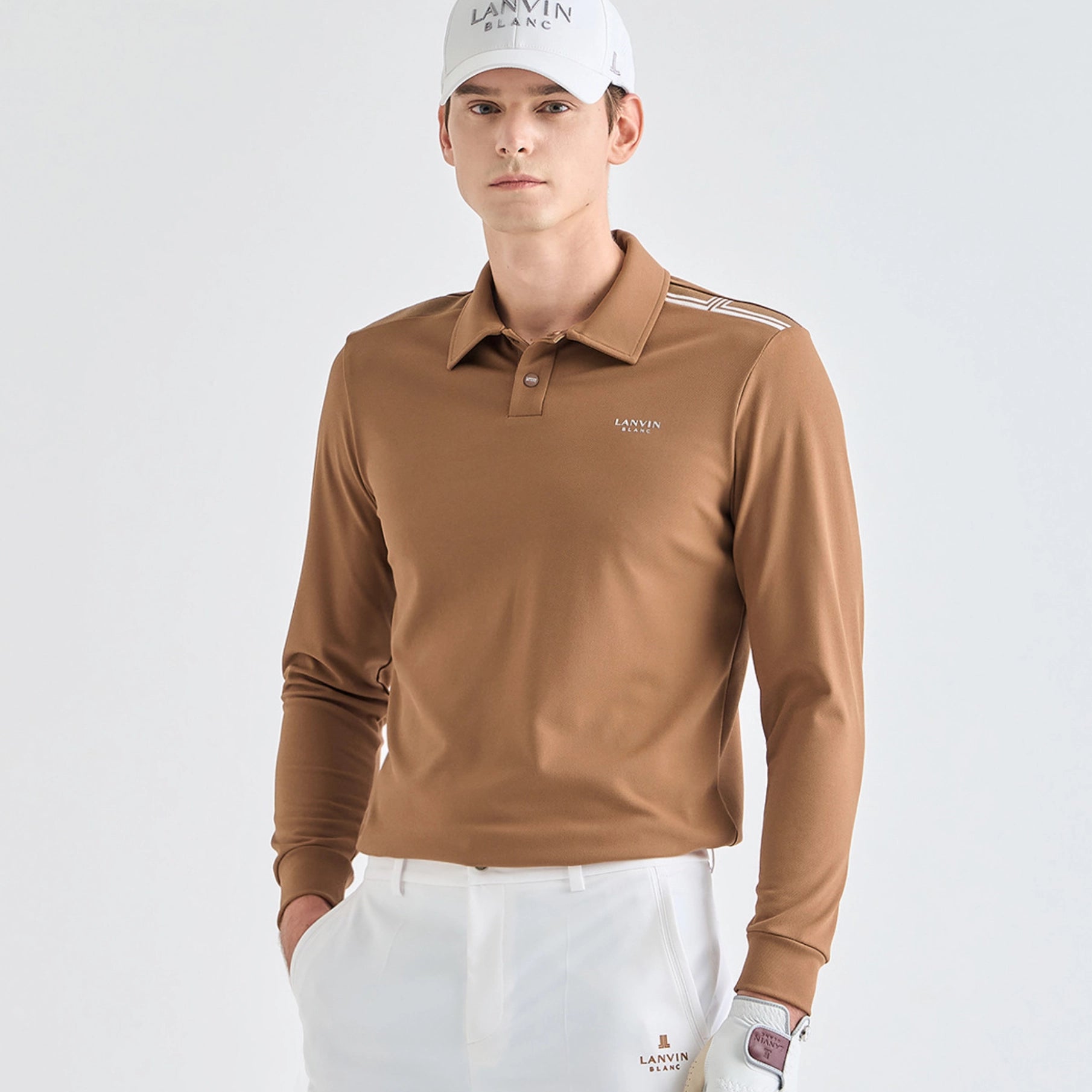 A male model wearing a tan long-sleeve golf polo shirt with white pants and a white cap, standing against a light gray studio background.