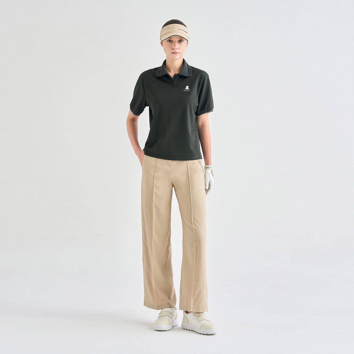 A woman modeling a dark green short-sleeve polo shirt with a small embroidered logo on the left chest, paired with beige pleated wide-leg trousers, a beige visor, a white golf glove, and white golf shoes, against a plain light gray studio background.