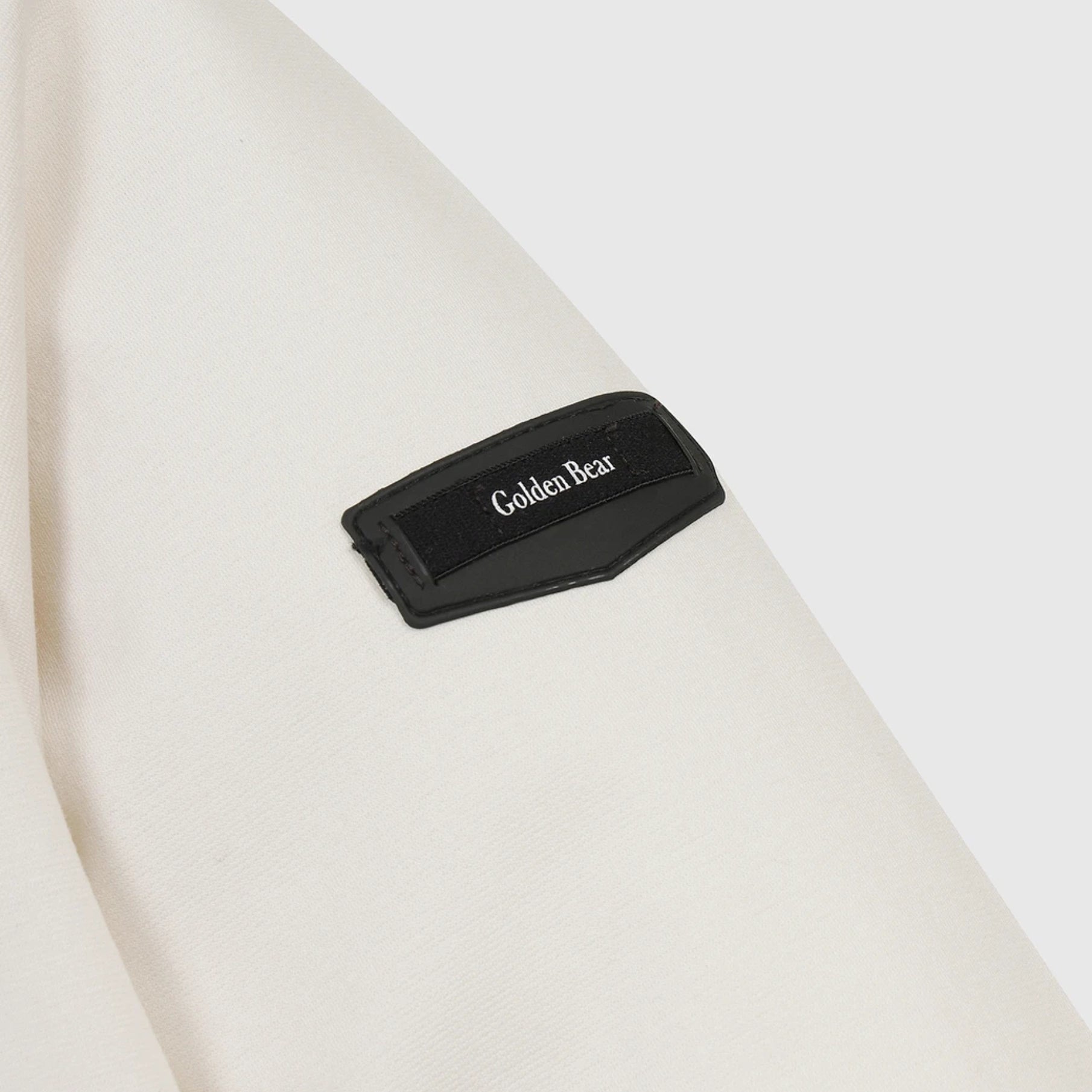 A close-up of a black fabric patch with the brand name 'Golden Bear' embroidered in white, attached to a light cream-colored textured garment.