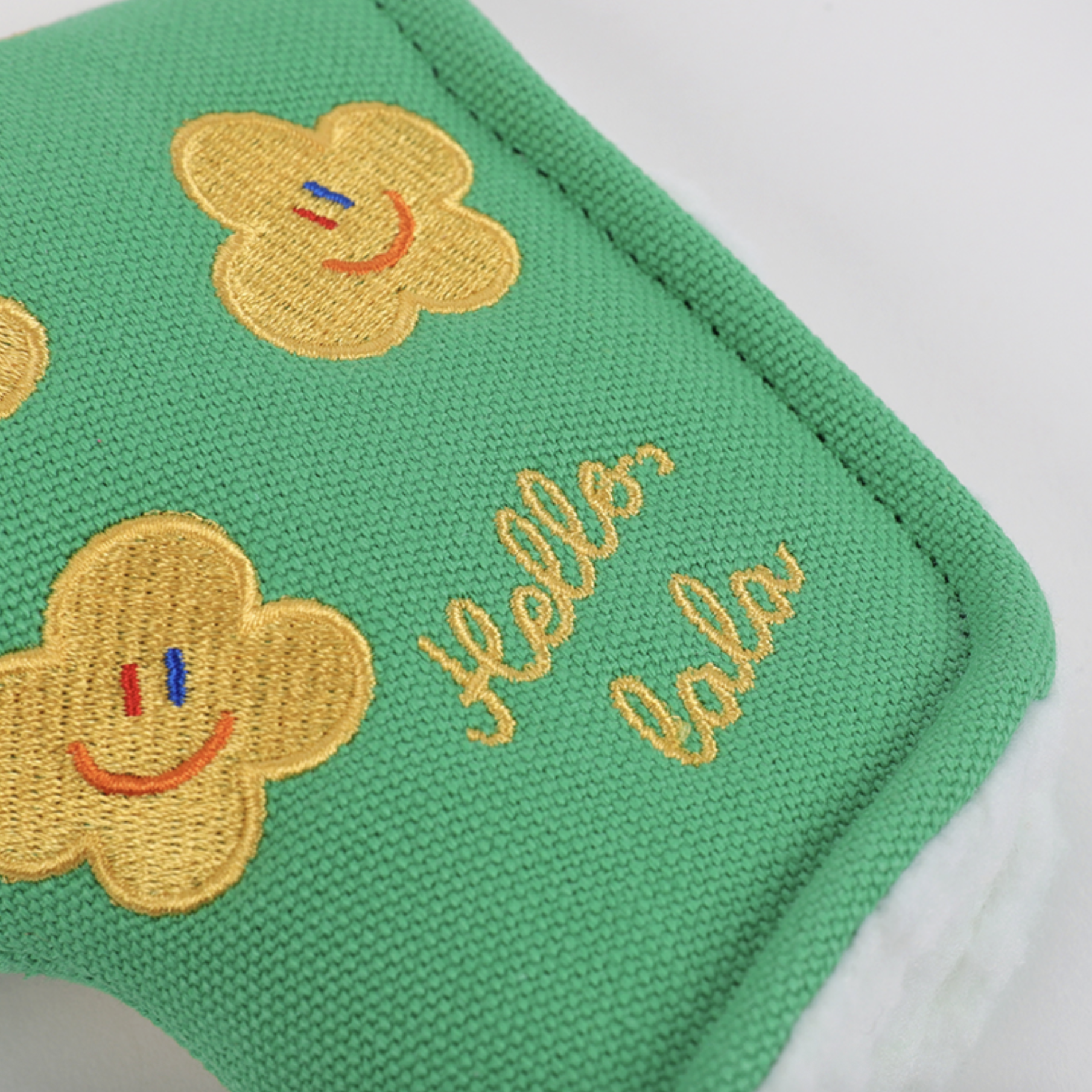 Close-up of a green woven fabric item with embroidered yellow smiley-face flowers and gold script reading 'hello lala' on a white background.