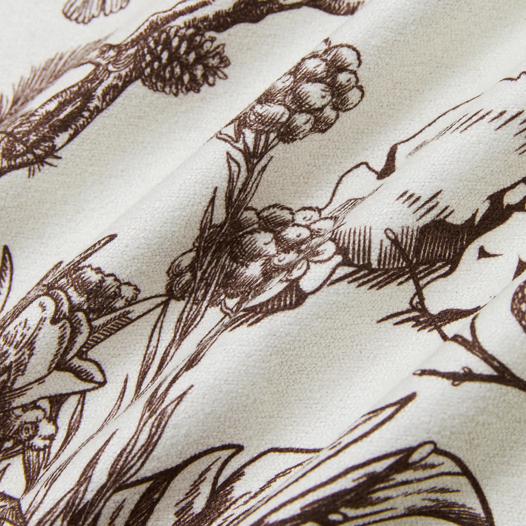 Close-up of a cream-colored fabric with a dark brown botanical and bird illustration pattern.