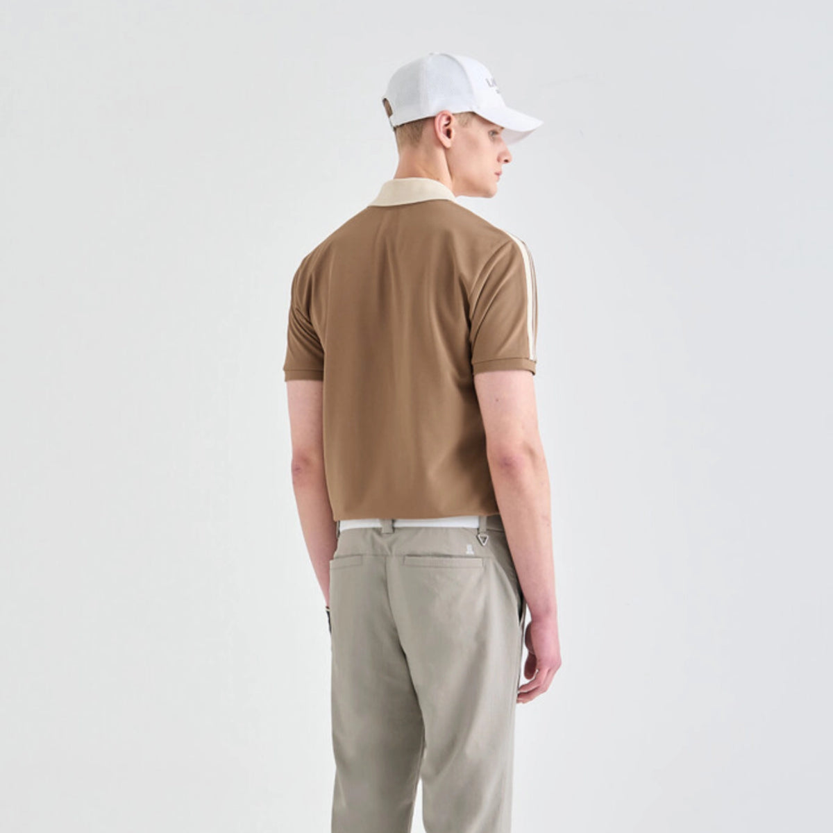 A man wearing a brown short-sleeve polo shirt with a cream collar and white sleeve stripes, paired with beige trousers and a white baseball cap, shown from the back against a light gray studio background.