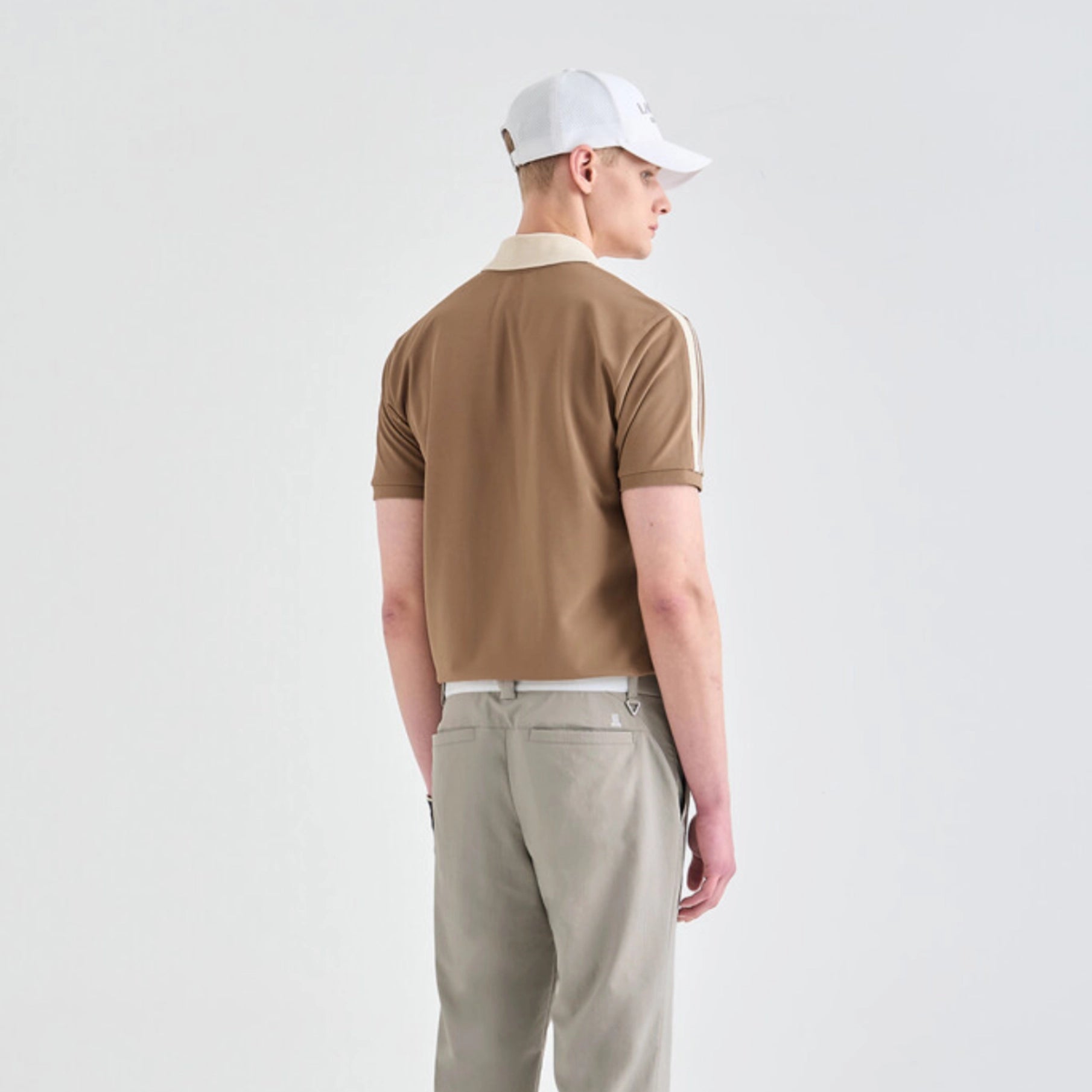 A man wearing a brown short-sleeve polo shirt with a cream collar and white sleeve stripes, paired with beige trousers and a white baseball cap, shown from the back against a light gray studio background.