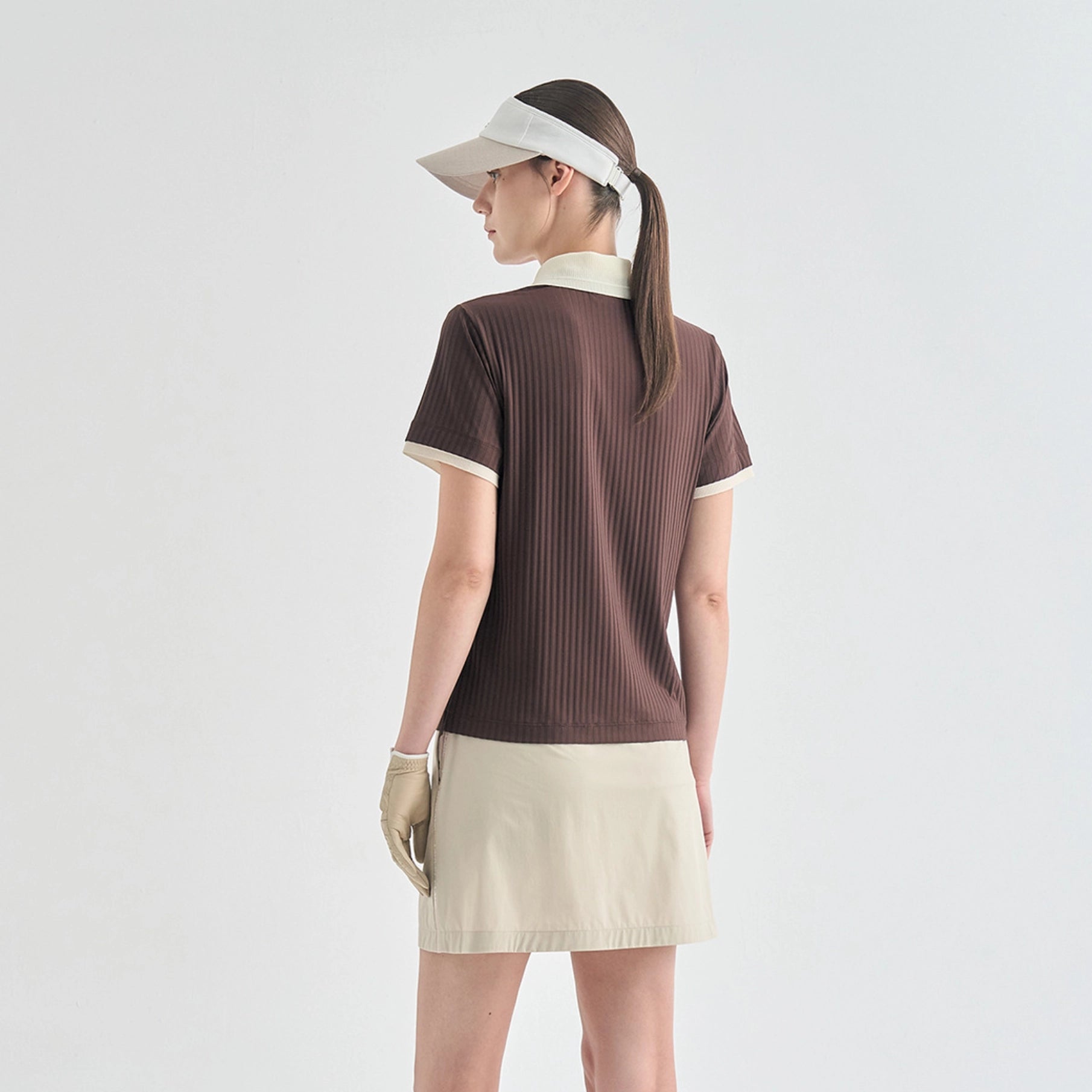A woman shown from the back wearing a brown ribbed short-sleeve polo shirt with cream trim, a cream skirt, a white visor, and a beige glove, against a plain light gray studio background.