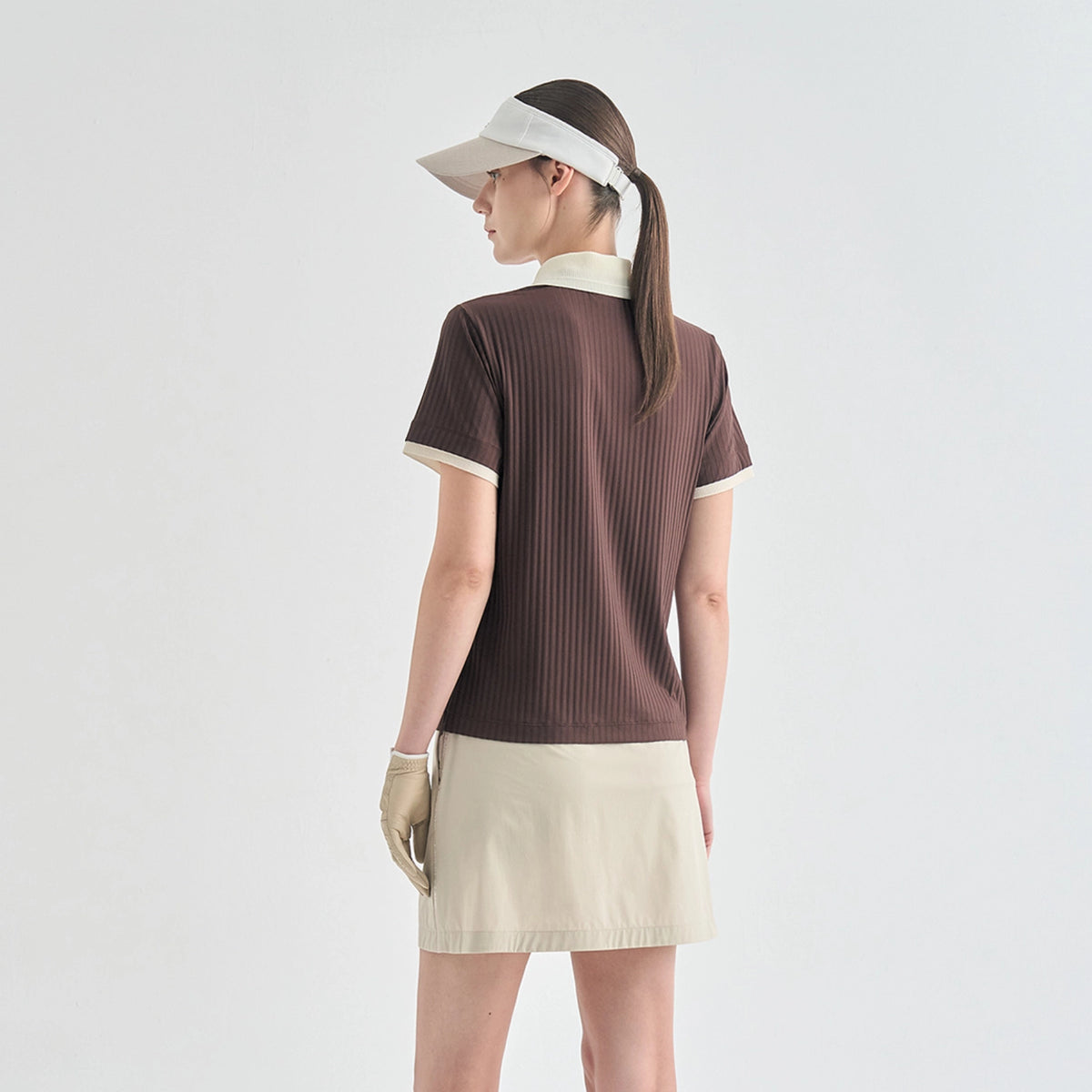 A woman shown from the back wearing a brown ribbed short-sleeve polo shirt with cream trim, a cream skirt, a white visor, and a beige glove, against a plain light gray studio background.