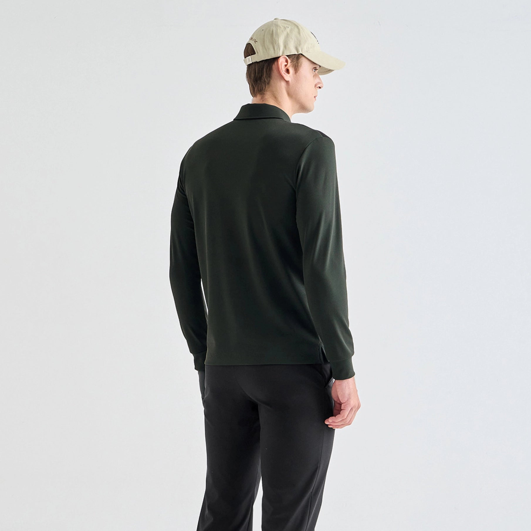 A man wearing a dark forest green long-sleeve polo shirt, black pants, and a beige baseball cap, shown from a rear three-quarter angle against a light gray studio background.