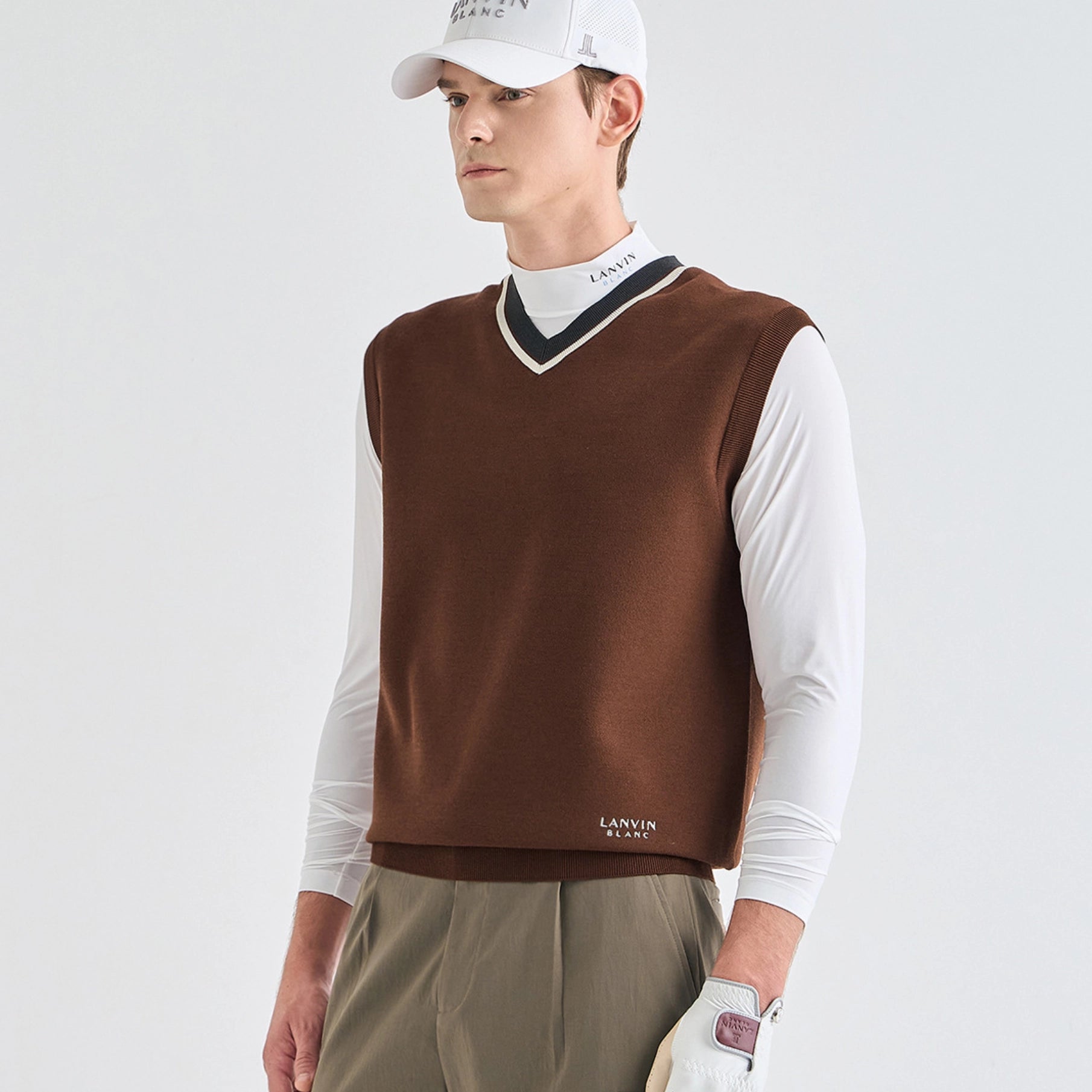 A male model wearing a brown knit V-neck sweater vest over a white long-sleeve collared shirt, paired with khaki pants, a white cap, and a golf glove. The brand name LANVIN is visible on the chest of the vest and on the cap.