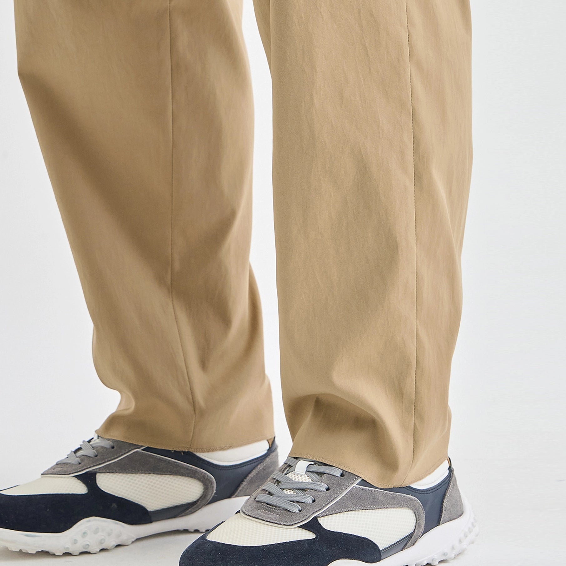 Close-up of a person's legs wearing beige trousers and navy, gray, and white sneakers against a plain light background.