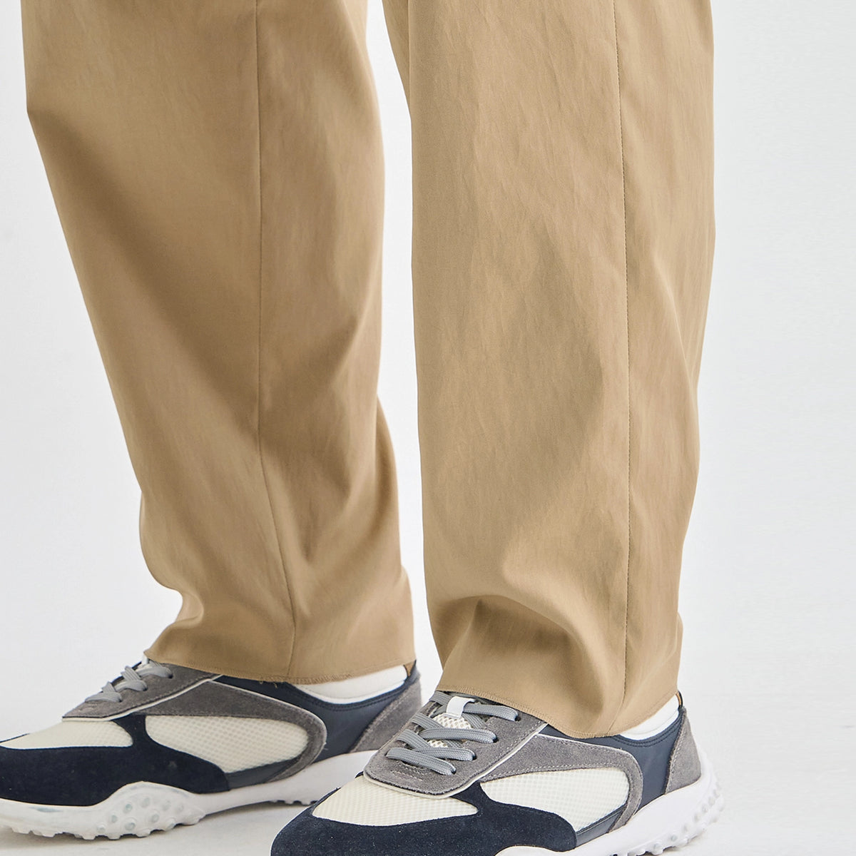 Close-up of a person's legs wearing beige trousers and navy, gray, and white sneakers against a plain light background.