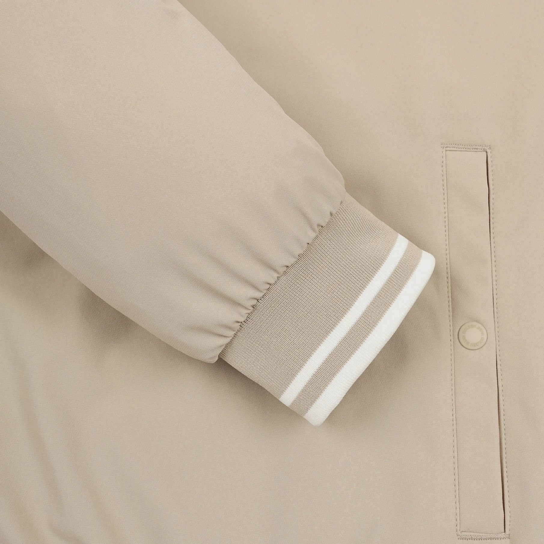 Close-up of a beige jacket sleeve with a ribbed cuff featuring two white stripes, next to a vertical welt pocket with a snap button.