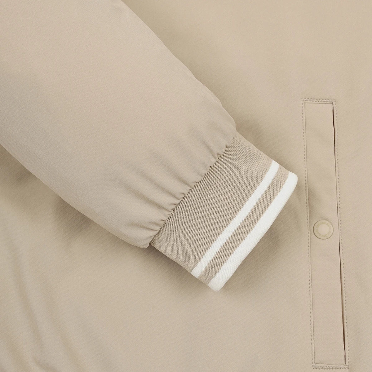 Close-up of a beige jacket sleeve with a ribbed cuff featuring two white stripes, next to a vertical welt pocket with a snap button.
