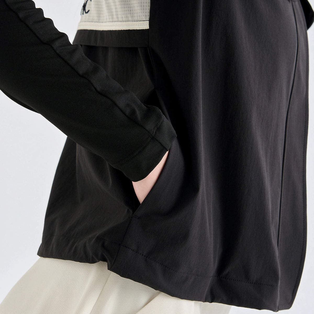 Close-up of a person wearing a black long-sleeve lightweight jacket with a side pocket, paired with a cream-colored top and skirt. The jacket has a subtle textured panel on the upper chest.