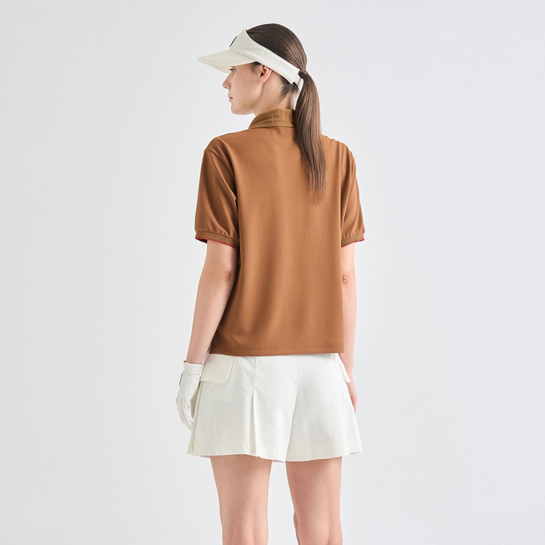 A woman shown from the back wearing a brown short-sleeve mock-neck athletic top, white tennis skirt, white visor, and white glove, against a plain light gray studio background.