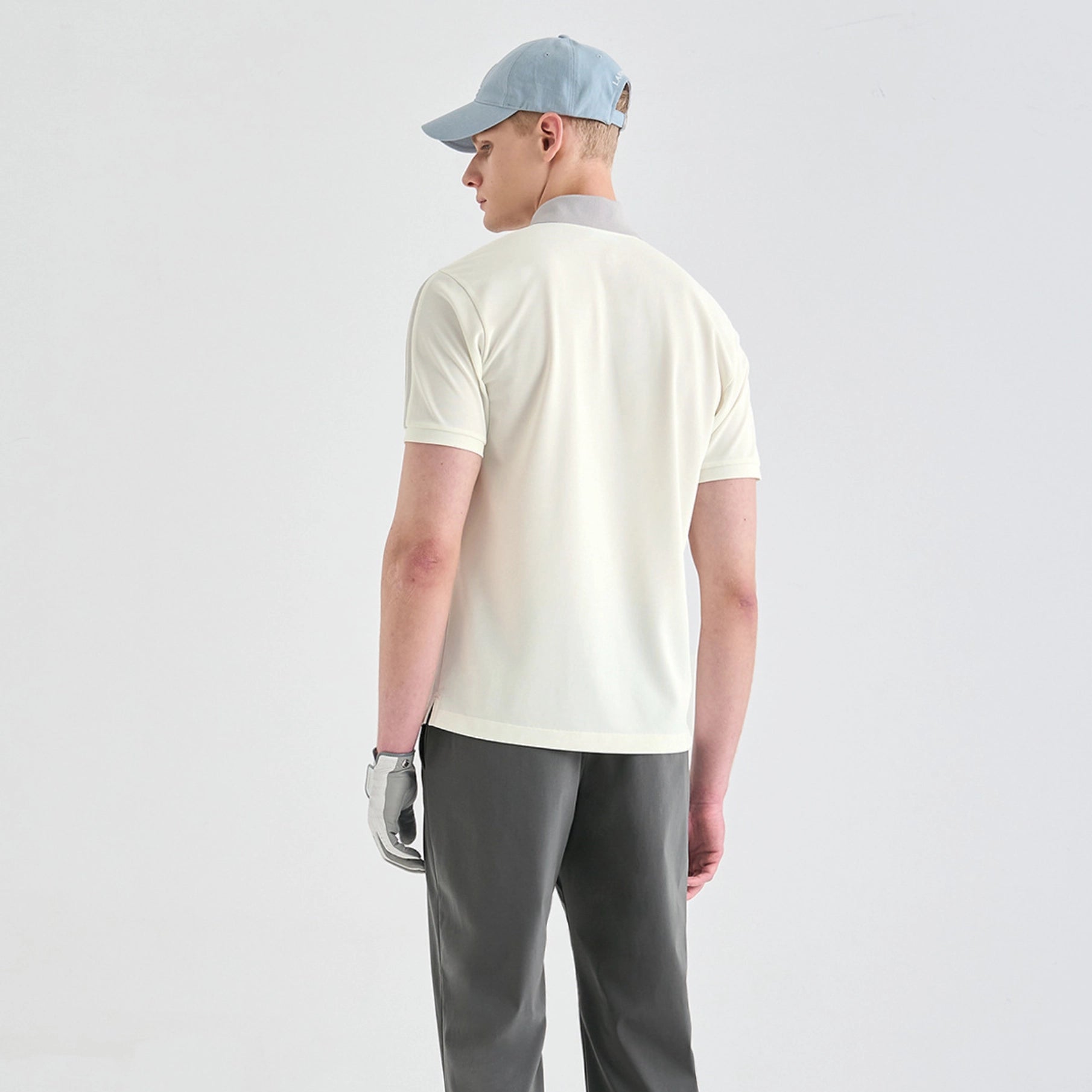 A man shown from the back wearing a light cream short-sleeve polo shirt, gray trousers, a light blue baseball cap, and a silver golf glove, against a plain light gray studio background.