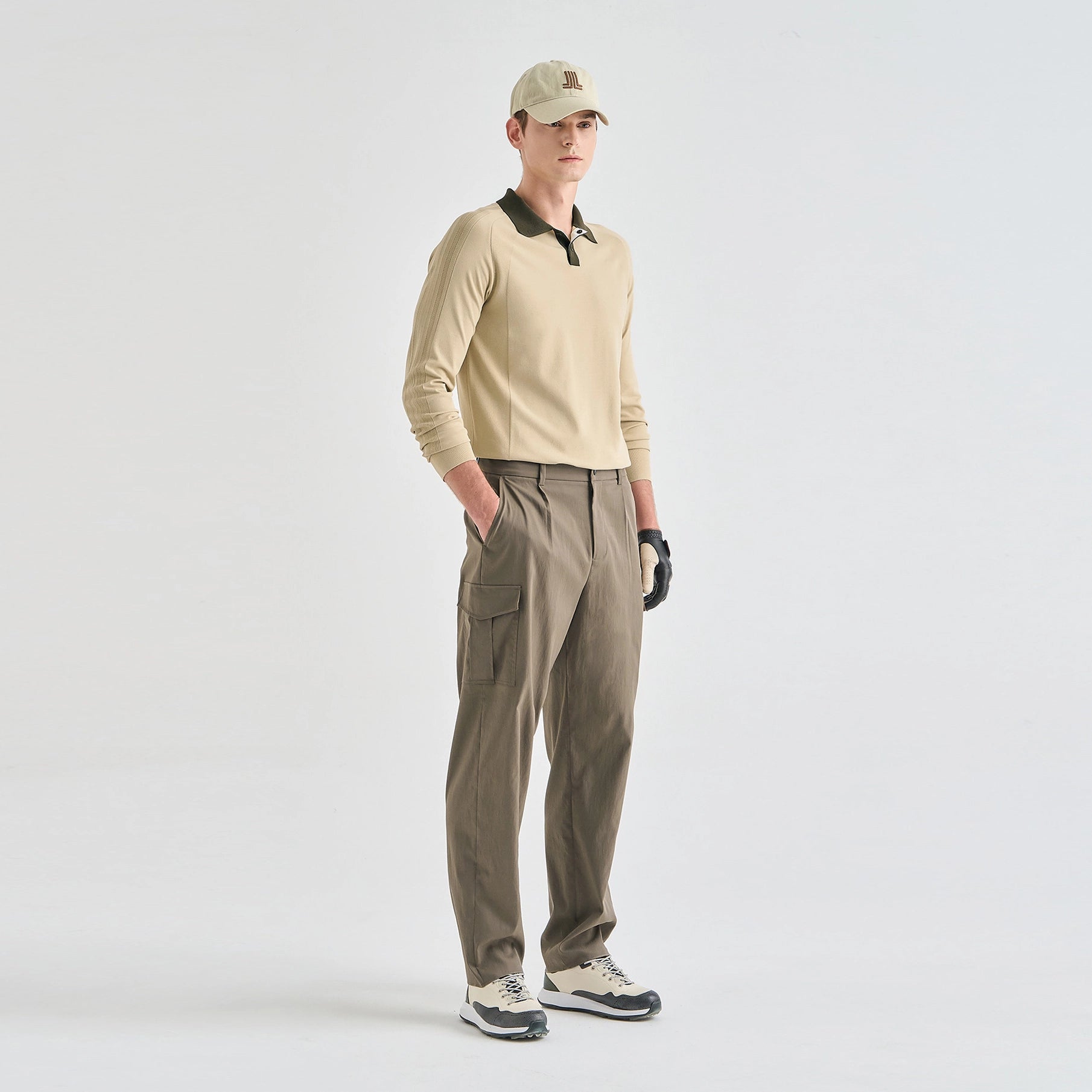 A male model stands against a light gray studio background wearing a beige long-sleeve polo shirt with a dark collar, olive cargo pants, a beige cap, black gloves, and white-and-black sneakers.