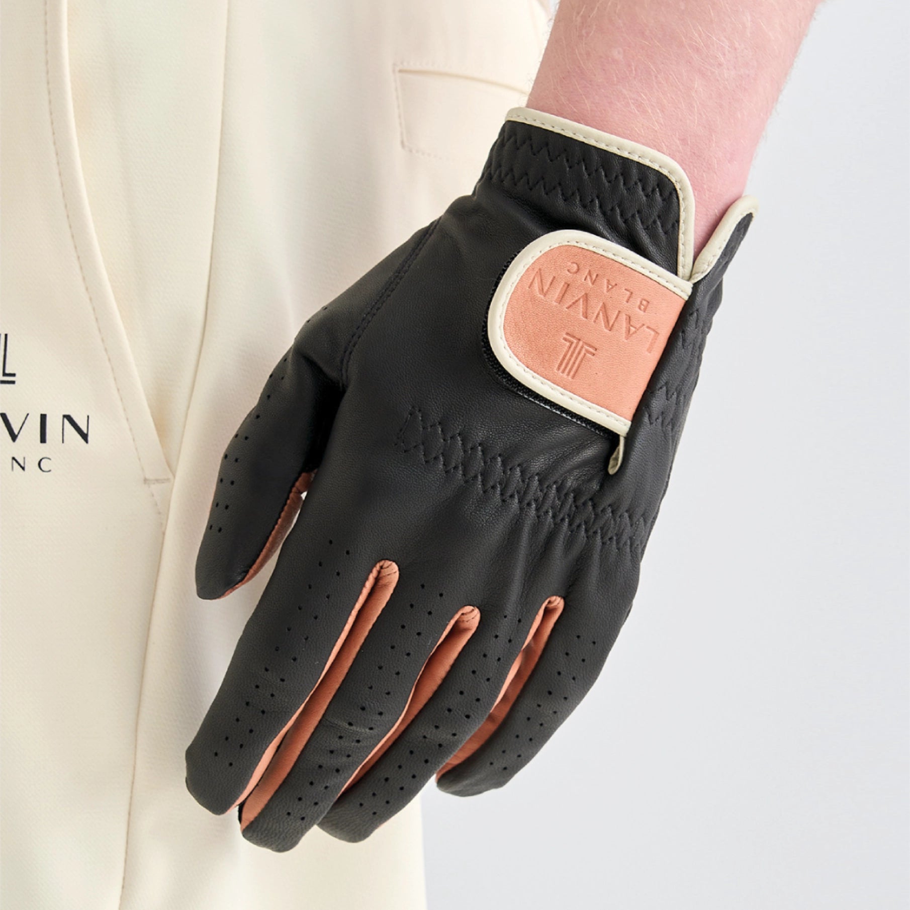 A close-up of a black leather golf glove with tan leather accents and a tan brand patch, worn on a model's hand against a light cream blazer.