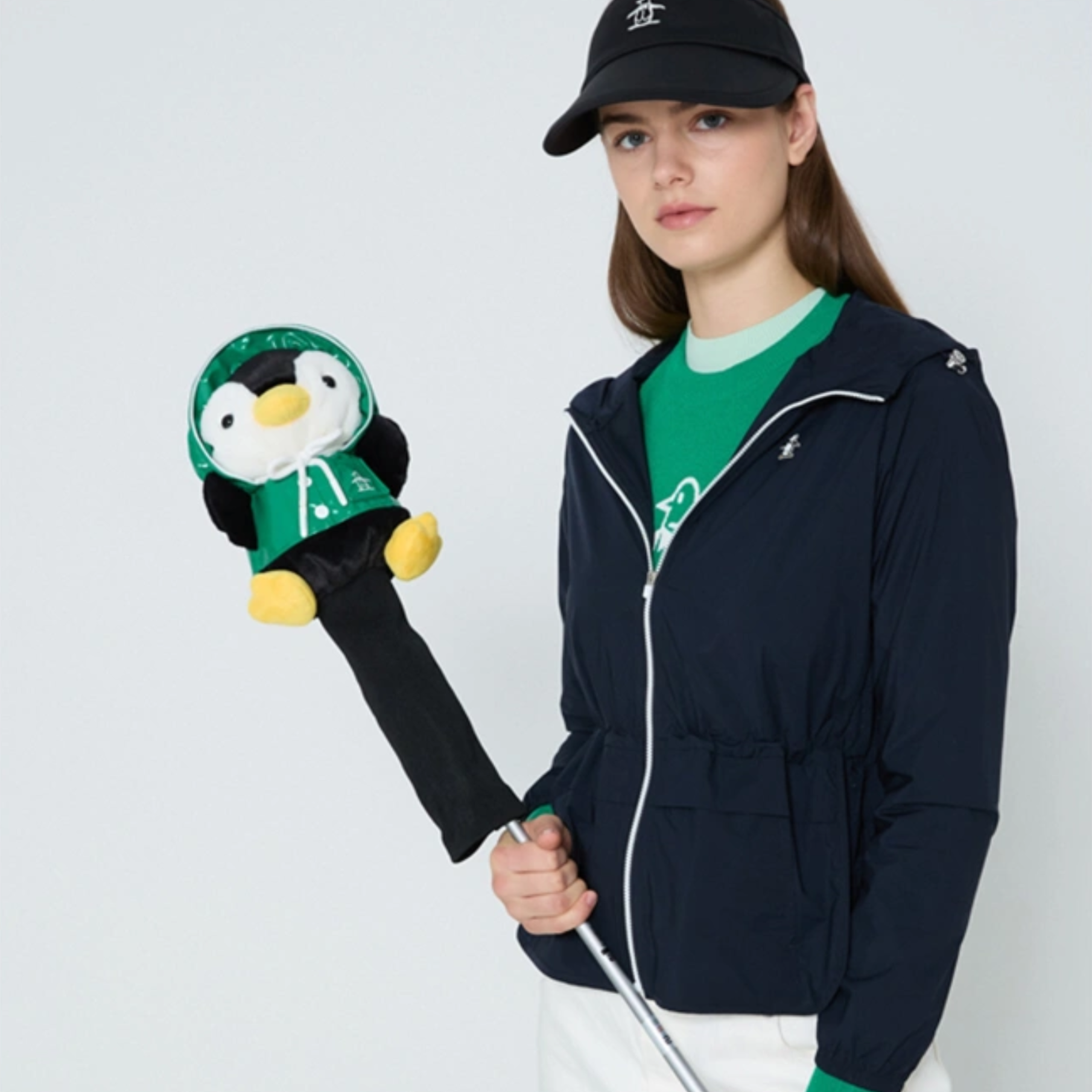 A woman wearing a navy zip-up jacket, green t-shirt, and black cap holds a golf club with a plush penguin headcover against a light gray studio background.