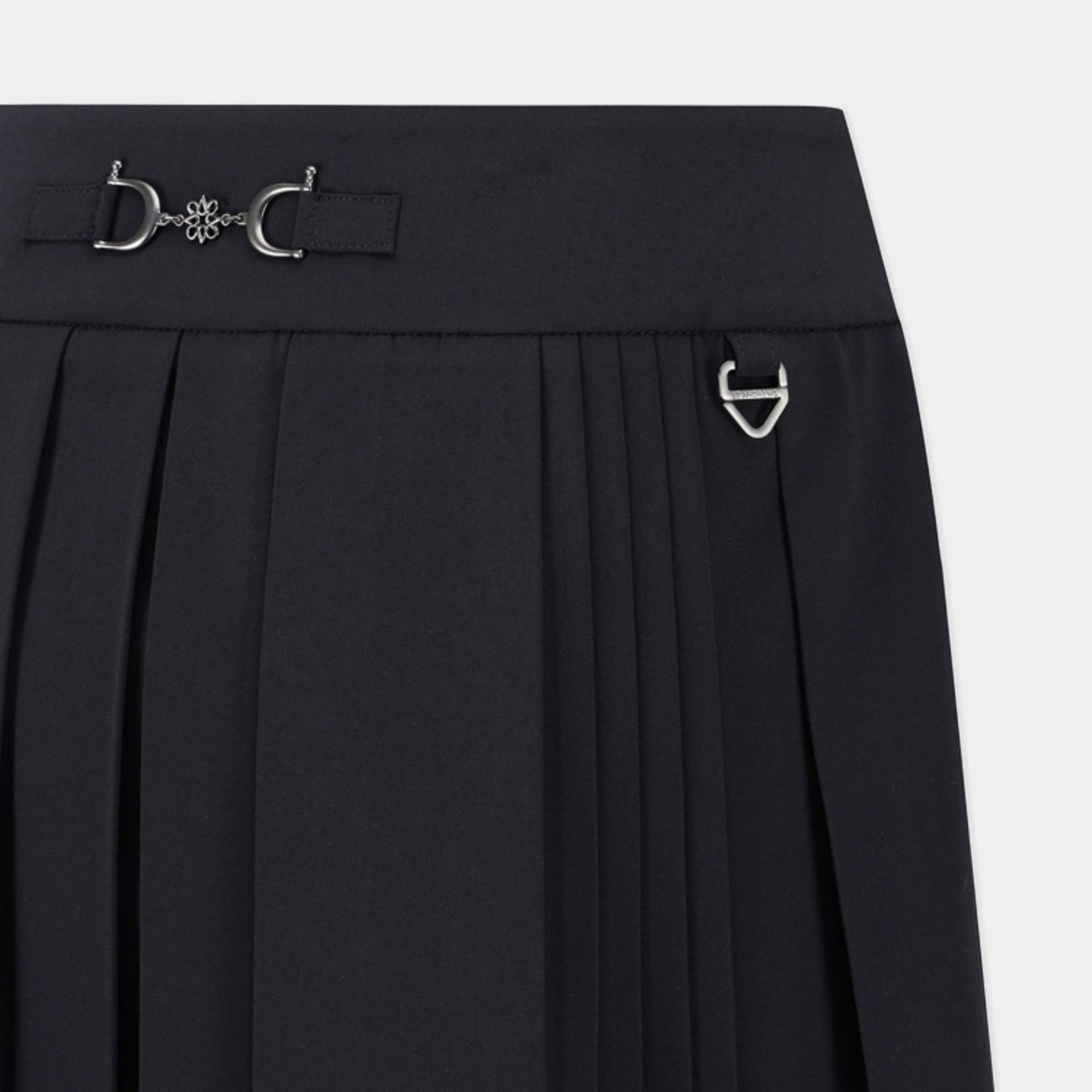 Close-up of a black pleated skirt with a wide waistband featuring silver-tone decorative hardware, including a horsebit-style detail and a small D-ring accent, on a light gray background.