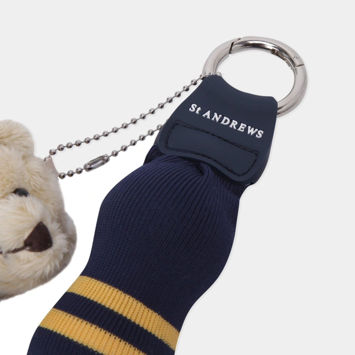 A St Andrews collegiate-themed keychain featuring a navy blue sock with yellow stripes, a navy blue strap with white embroidered 'ST ANDREWS' text, a silver metal key ring, and a silver ball chain. A plush cream-colored teddy bear head is partially visible on the left side.