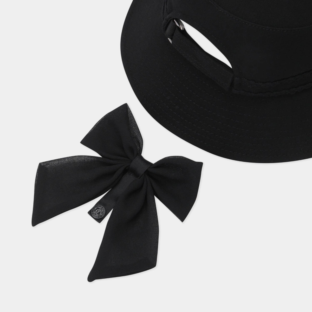 A black fabric bow accessory with a small black brand tag, displayed next to a cropped view of a black sun hat on a light gray background.