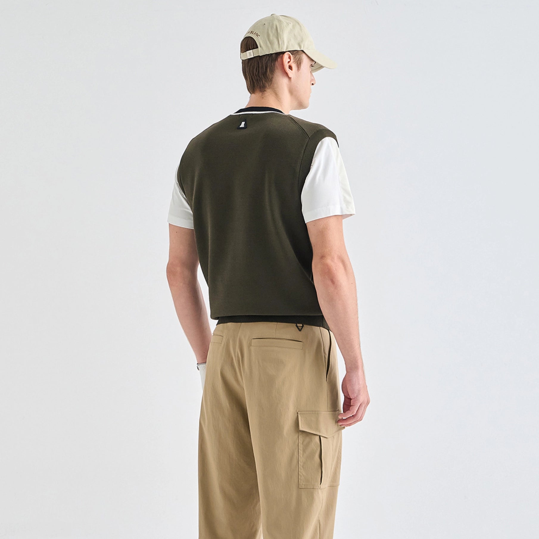 A man shown from the back wearing a dark olive green sleeveless knit vest over a white short-sleeve shirt, paired with beige cargo pants and a beige baseball cap, against a light gray studio background.