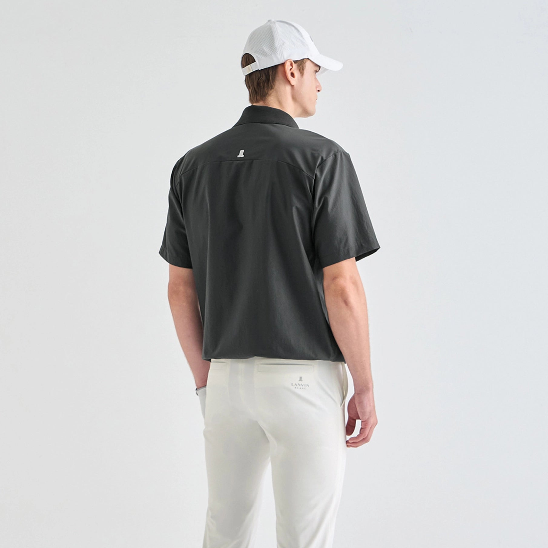 A man shown from the back wearing a dark charcoal gray short-sleeve collared shirt, white trousers, and a white baseball cap, against a plain light gray studio background.