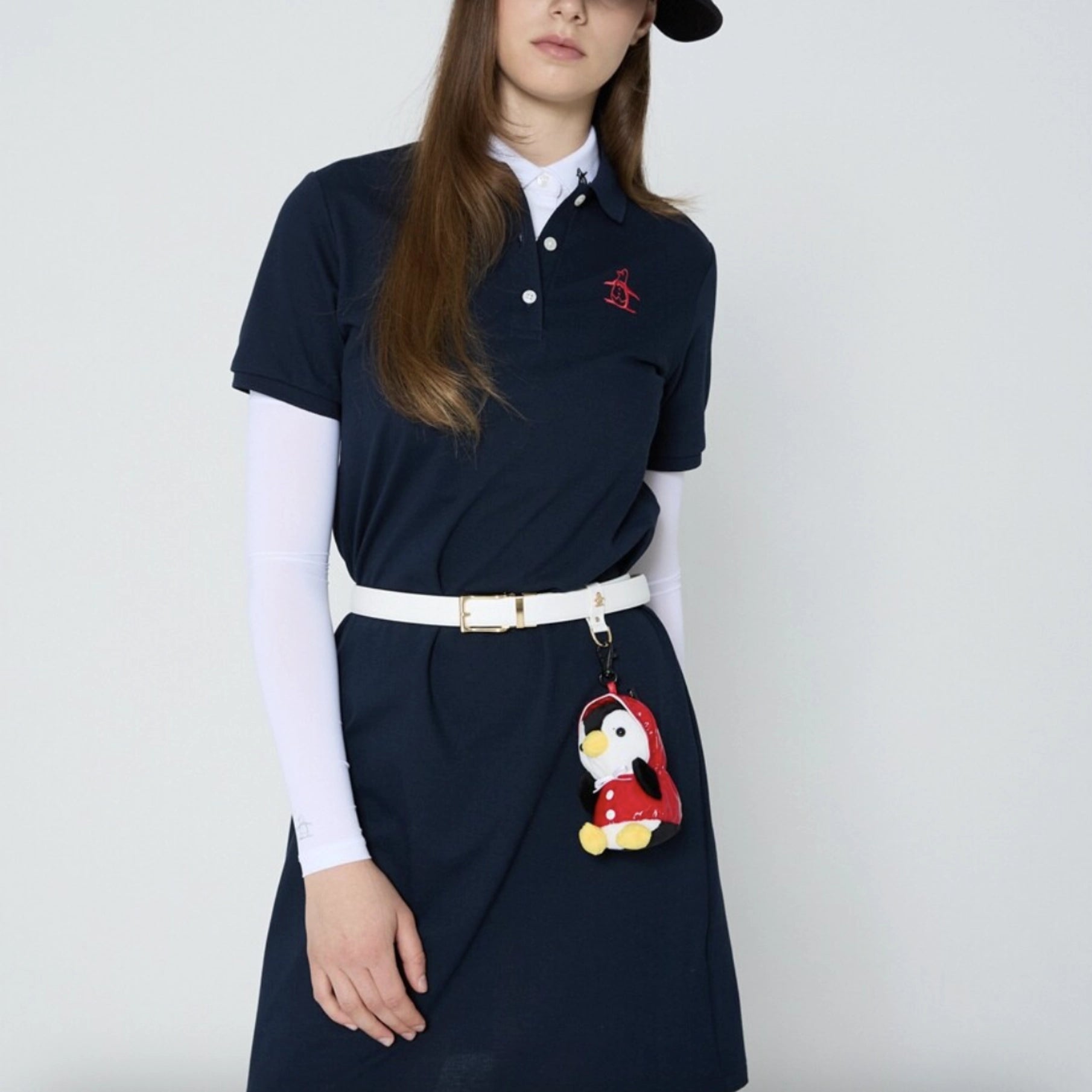 A model wearing a navy blue polo dress with a white collar, layered over a white long-sleeve shirt and a white belt. The polo dress features a small red embroidered logo on the left chest.