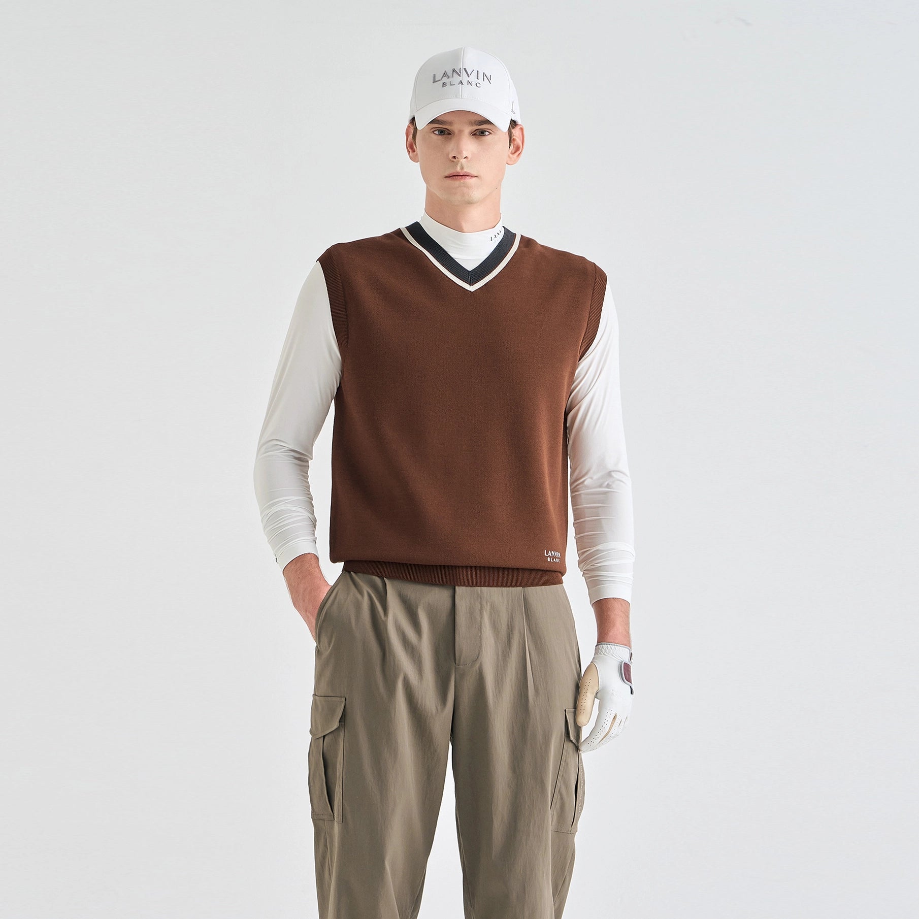A male model wearing a brown knit sweater vest over a white long-sleeve shirt, paired with khaki cargo pants, a white cap with 'LANVIN BLANC' text, and a white golf glove, against a light gray studio background.