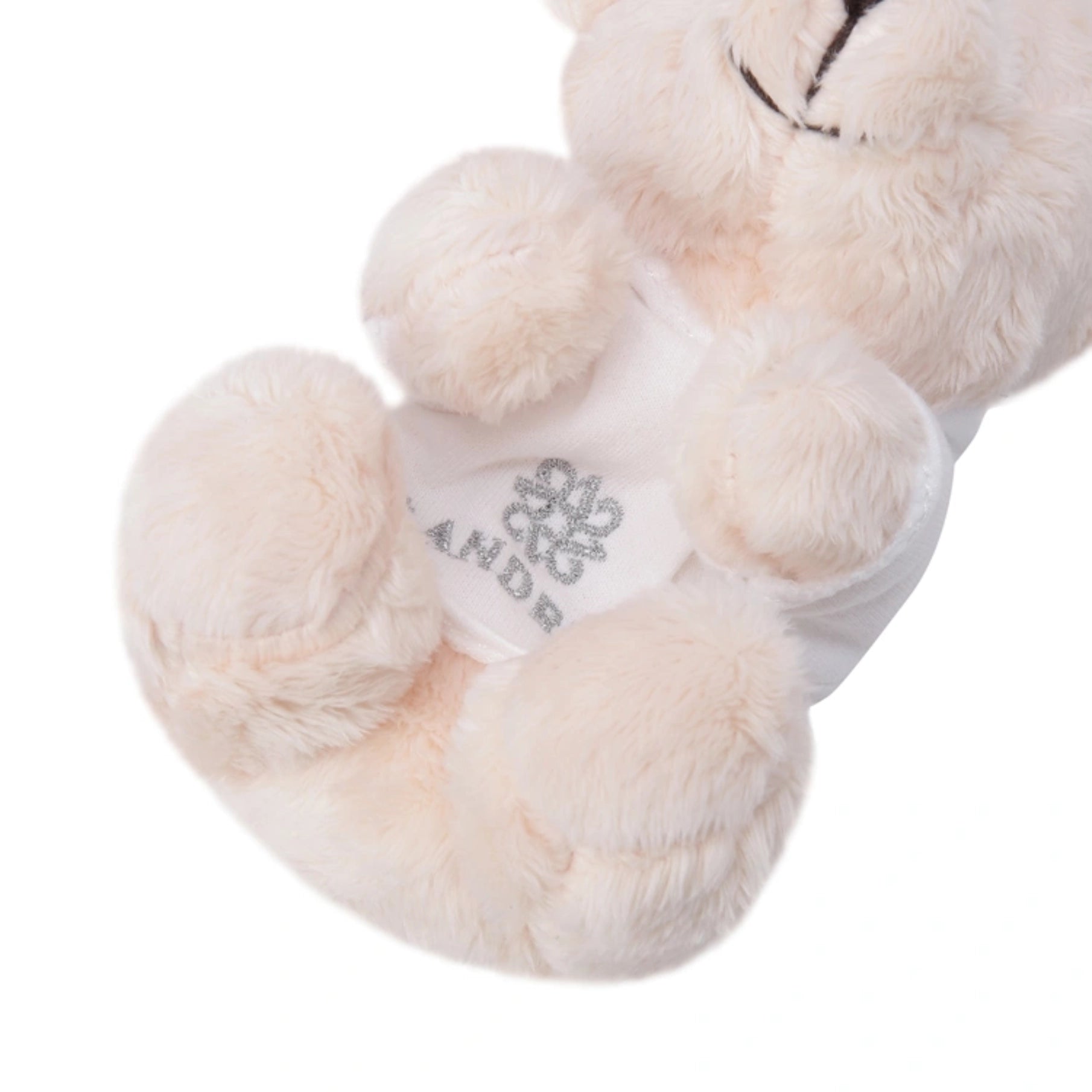 A close-up of a plush teddy bear in cream-colored faux fur, wearing a white shirt with a silver glittery Loewe anagram logo and the brand name 'ST ANDREWS'.