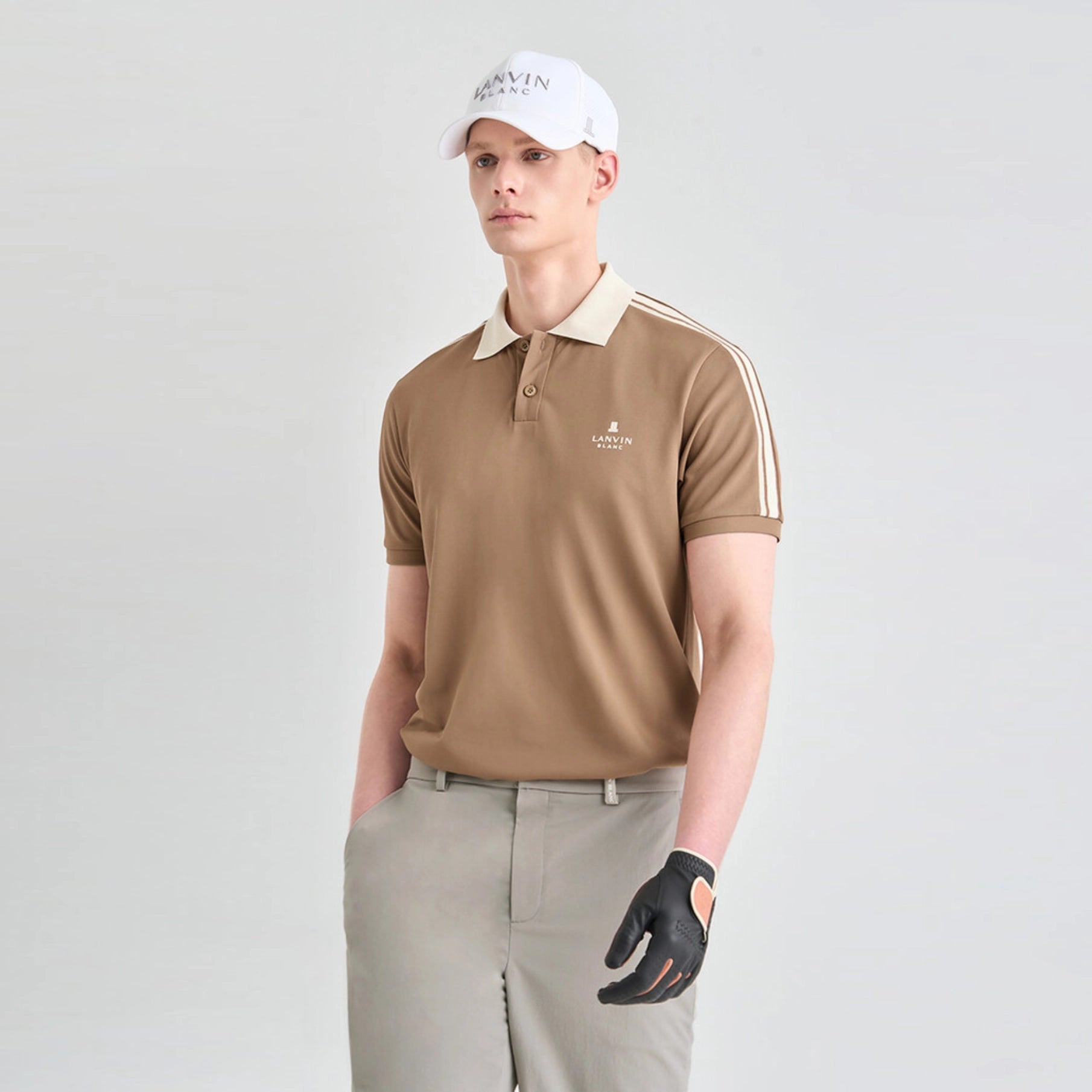 A man modeling a tan short-sleeve polo shirt with a beige collar and white stripes on the sleeves, paired with khaki pants, a white cap, and a black golf glove, against a light gray studio background.