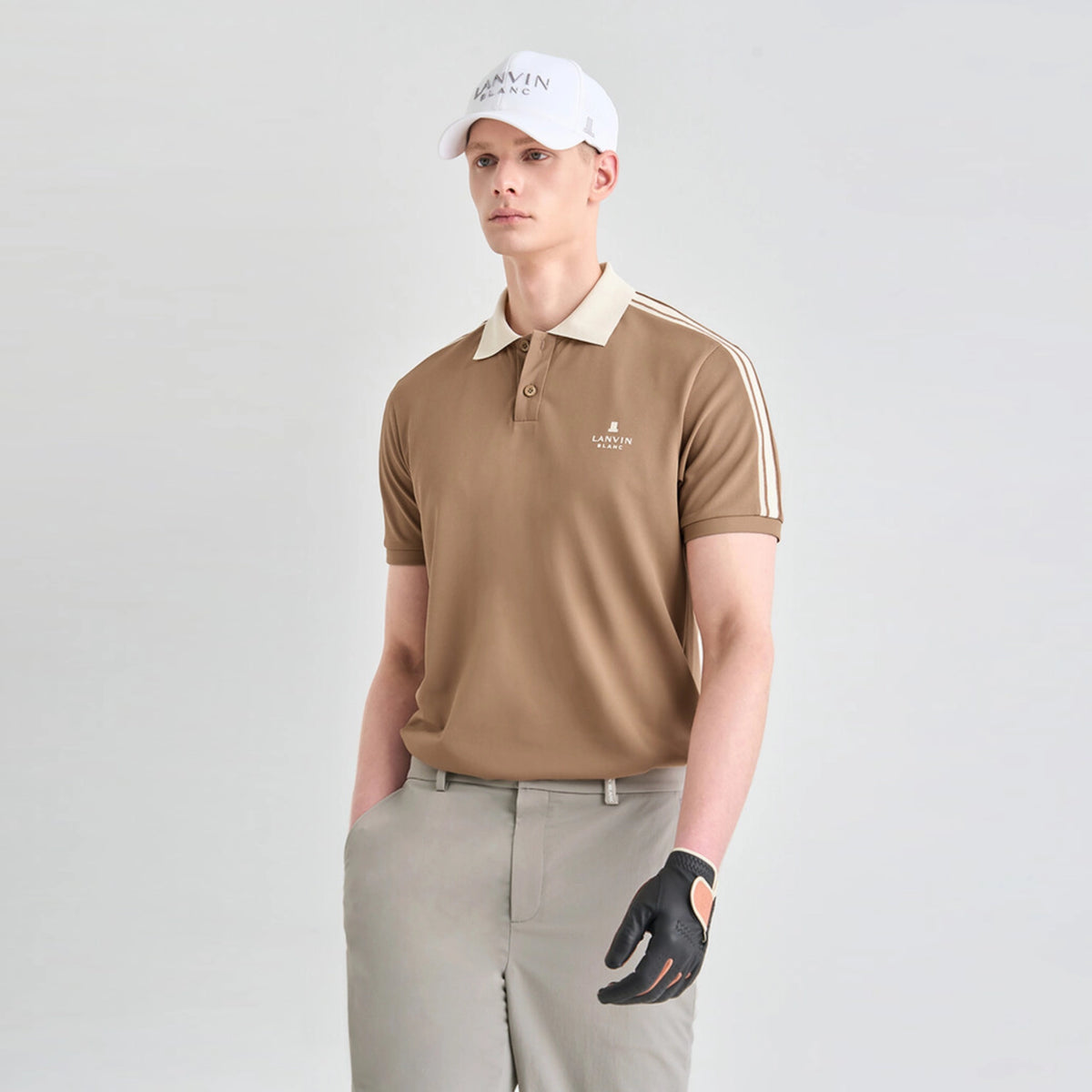 A man modeling a tan short-sleeve polo shirt with a beige collar and white stripes on the sleeves, paired with khaki pants, a white cap, and a black golf glove, against a light gray studio background.
