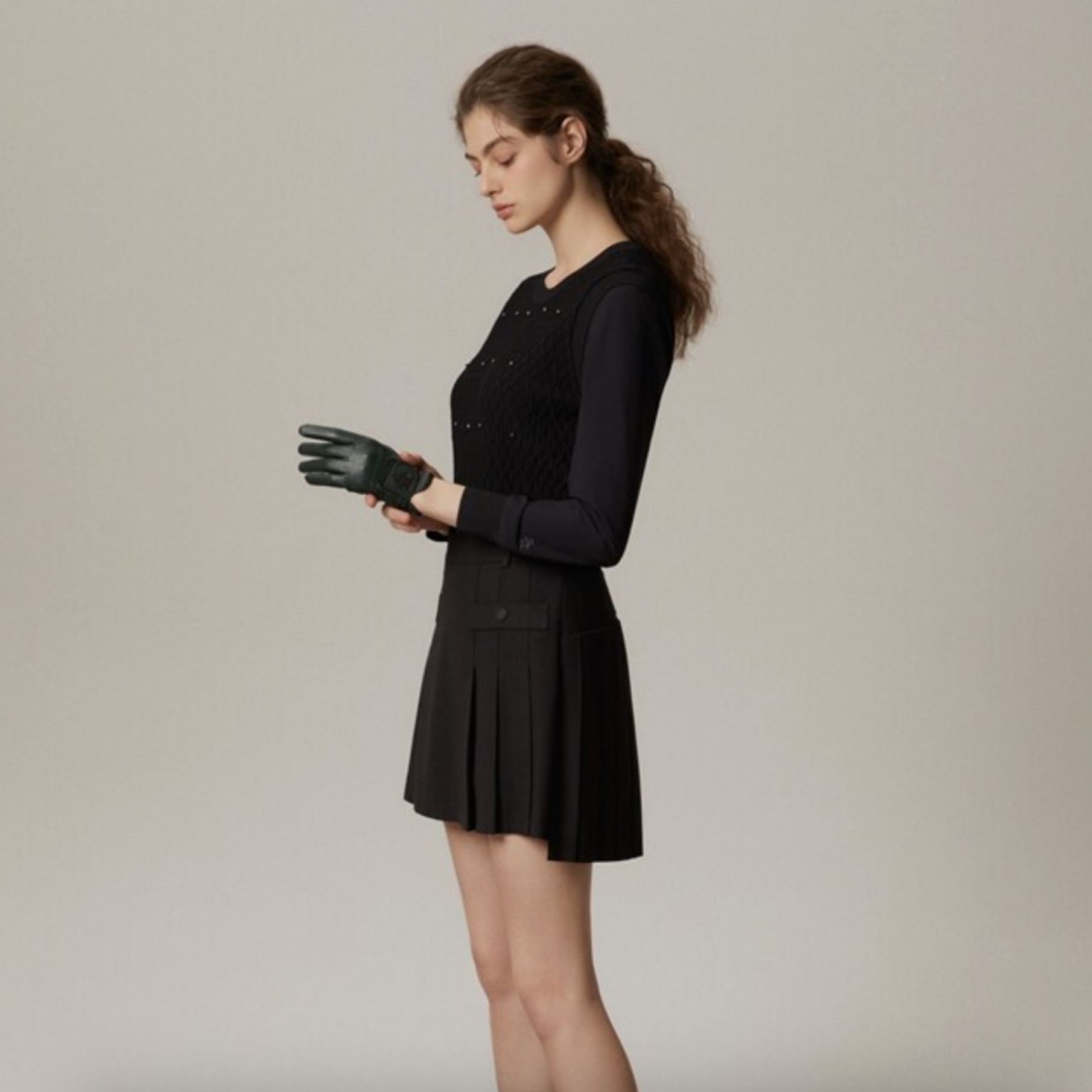 A woman wearing a black long-sleeve dress with a pleated skirt and black gloves, standing in profile against a plain light gray studio background.