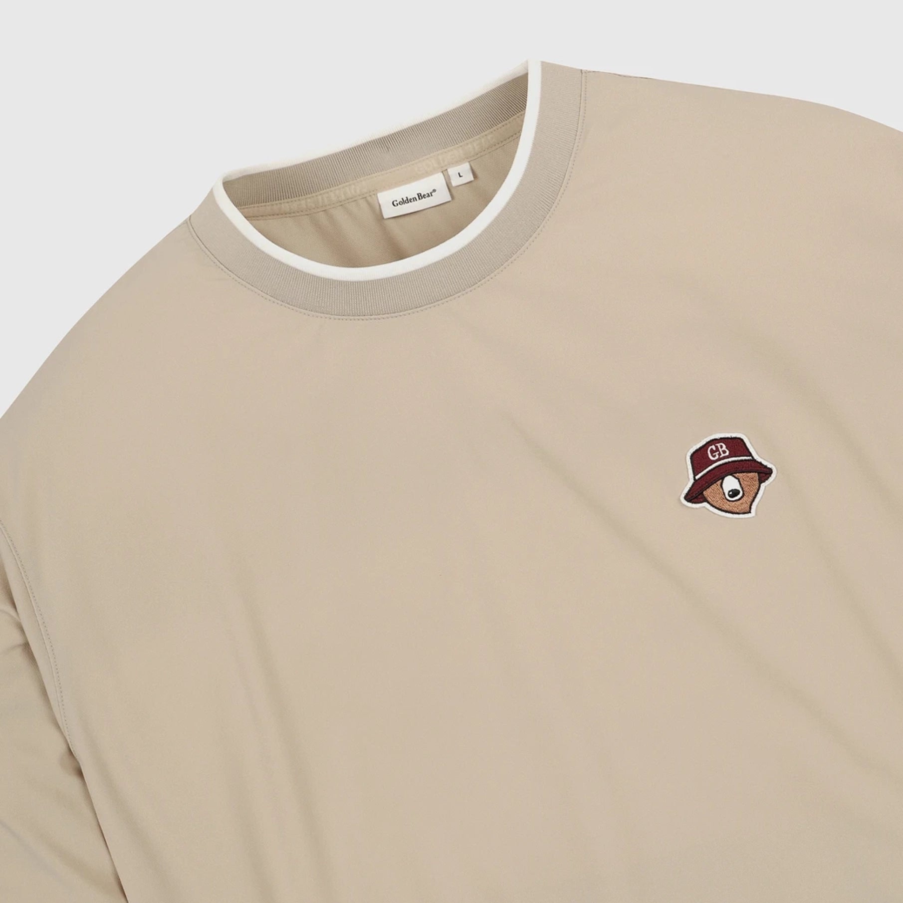 A close-up of a beige crewneck sweatshirt with a white ribbed collar trim and a small embroidered graphic on the left chest featuring a brown bear wearing a red hat.