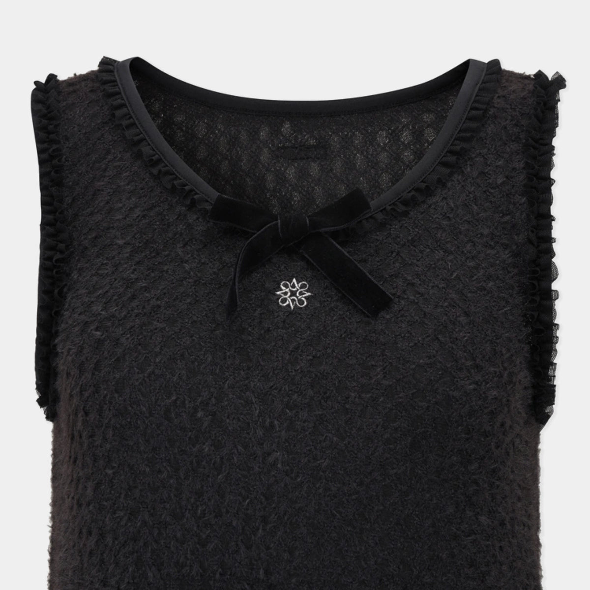 A close-up of a black textured sleeveless top with ruffled trim, a black bow at the neckline, and a small silver flower-shaped brooch on the chest, shown against a light gray background.