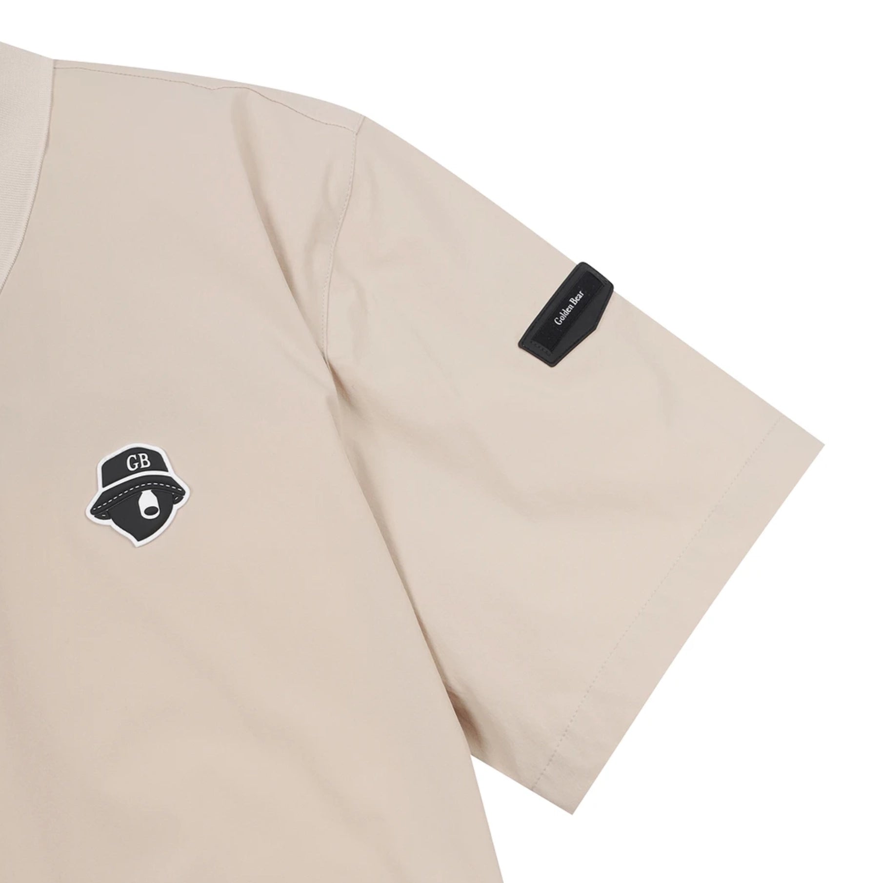 A close-up of a beige short-sleeve t-shirt showing the left chest with a black embroidered patch and the left sleeve with a black fabric brand tag.
