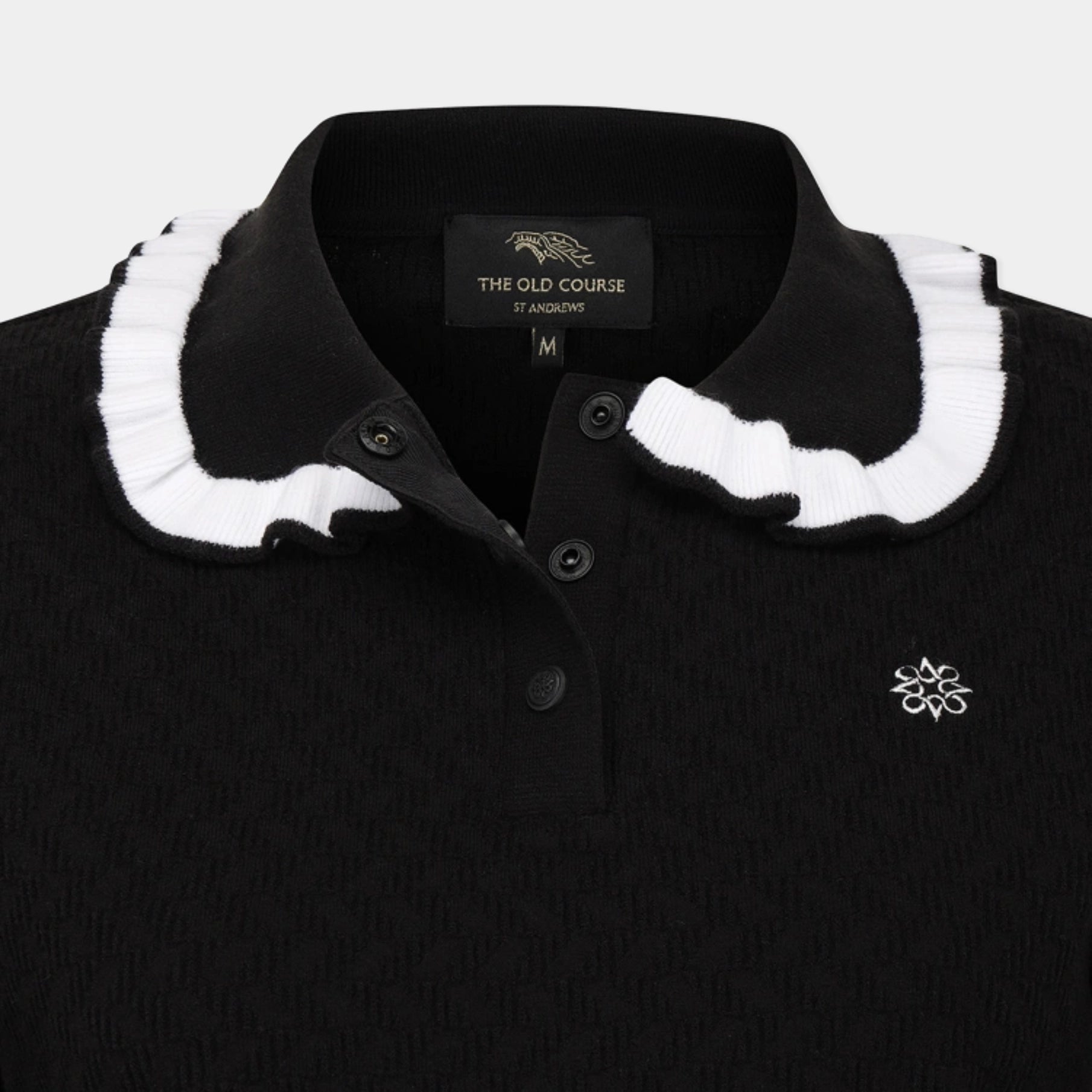 Close-up of a black polo shirt with a white ruffled collar, featuring a three-button placket and a small white embroidered logo on the chest. The inner neck label reads 'THE OLD COURSE' and 'ST ANDREWS'.