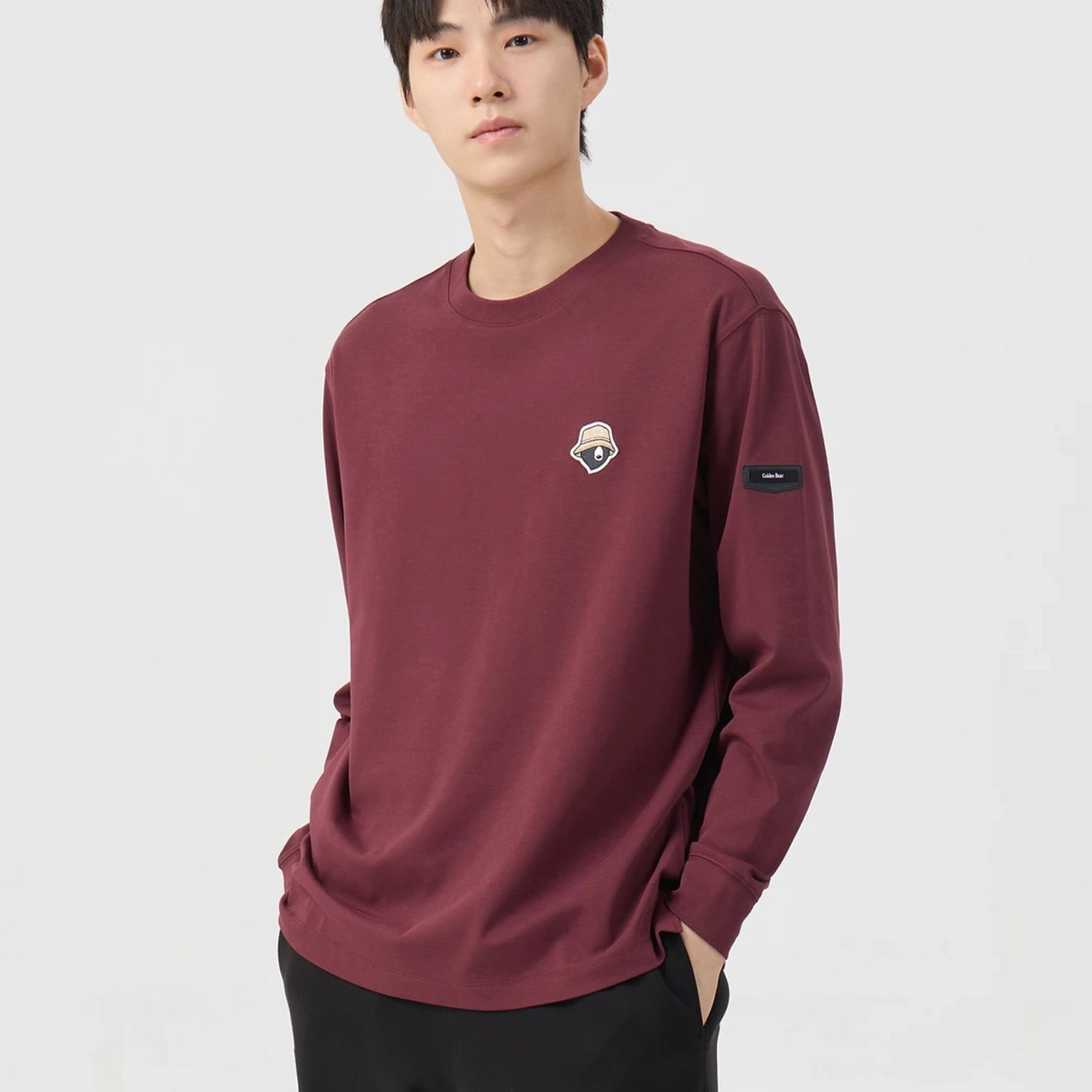 A man wearing a burgundy long-sleeve crewneck sweatshirt with a small shield logo on the left chest and a black patch on the right sleeve, set against a plain light gray background.