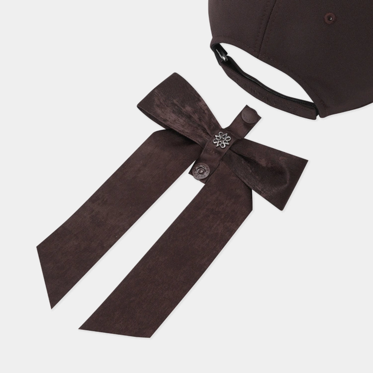 A dark brown cap with a long matching ribbon tail, featuring a bow with a small silver floral embellishment and buttons, shown on a light gray background.