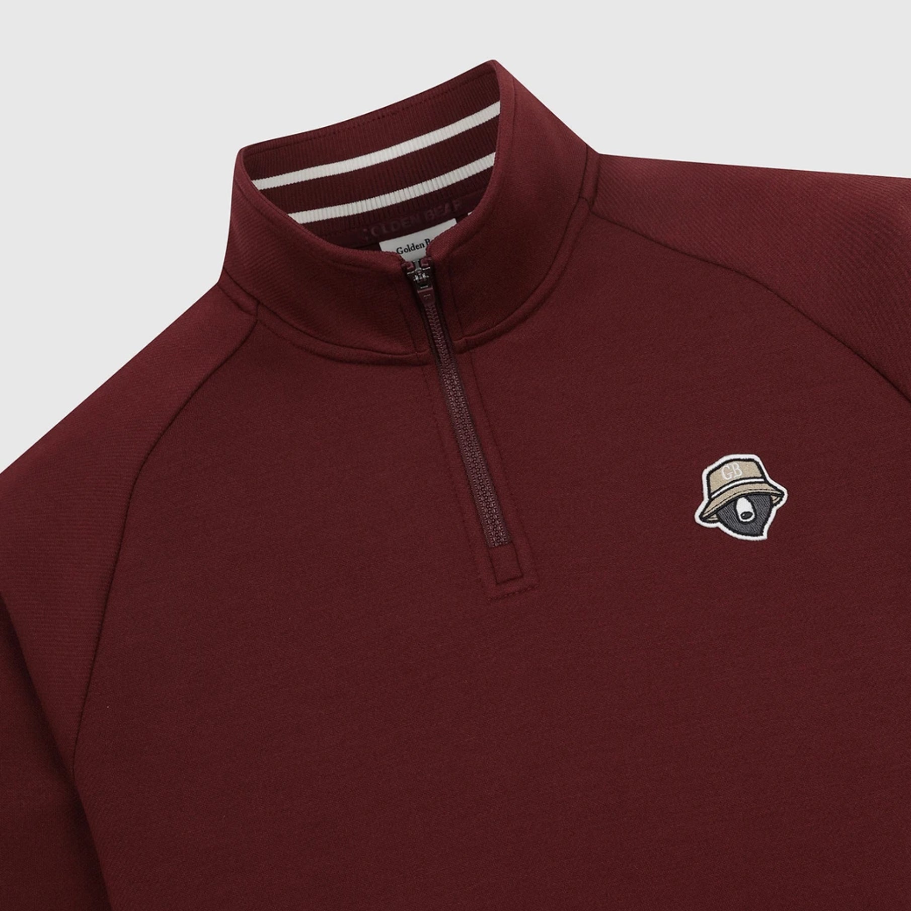 A close-up of a maroon quarter-zip pullover sweatshirt with a small embroidered hat logo on the left chest, set against a light gray background.