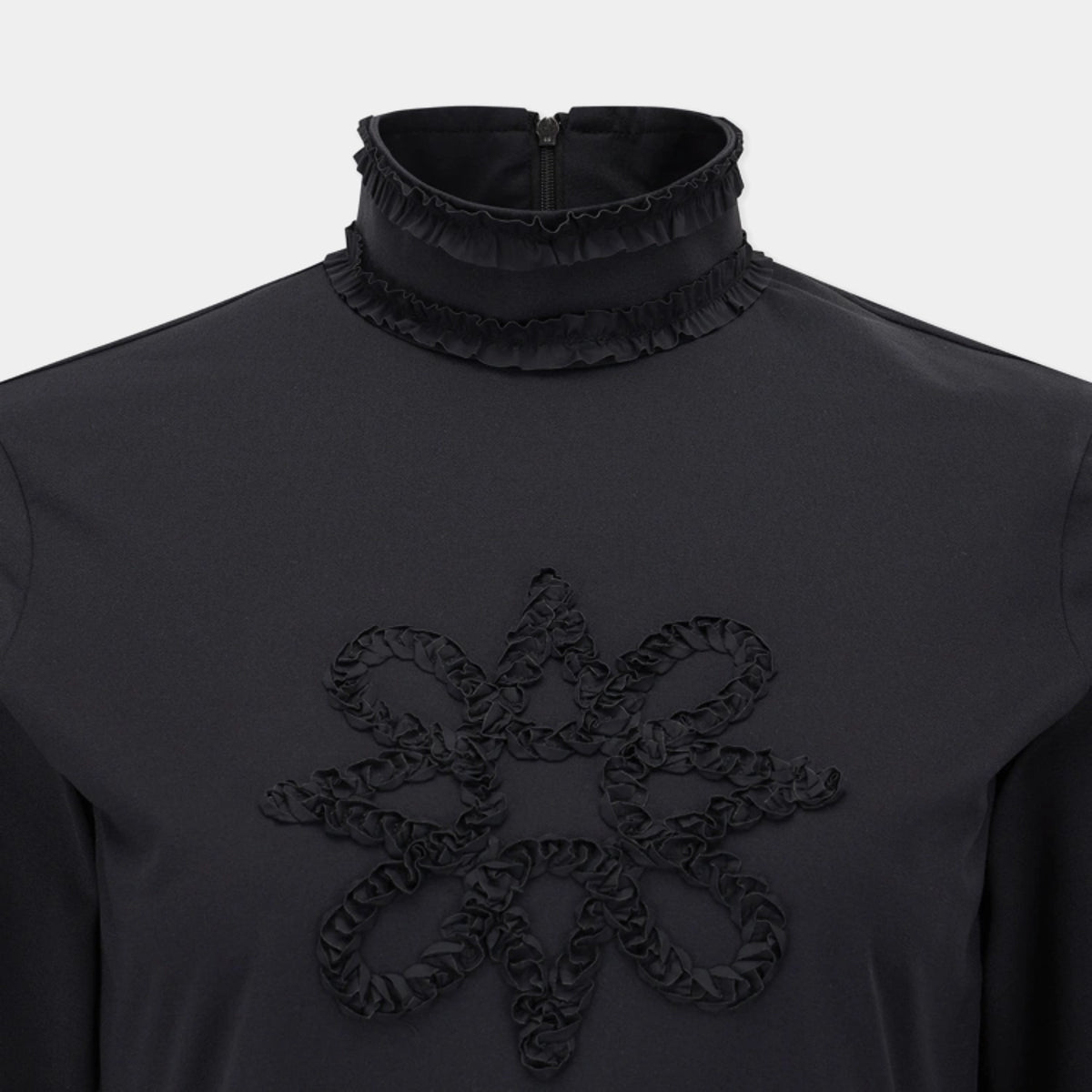 A close-up of a black long-sleeve top featuring a high ruffled mock neck and a large matching ruffled floral motif on the chest, set against a light gray background.