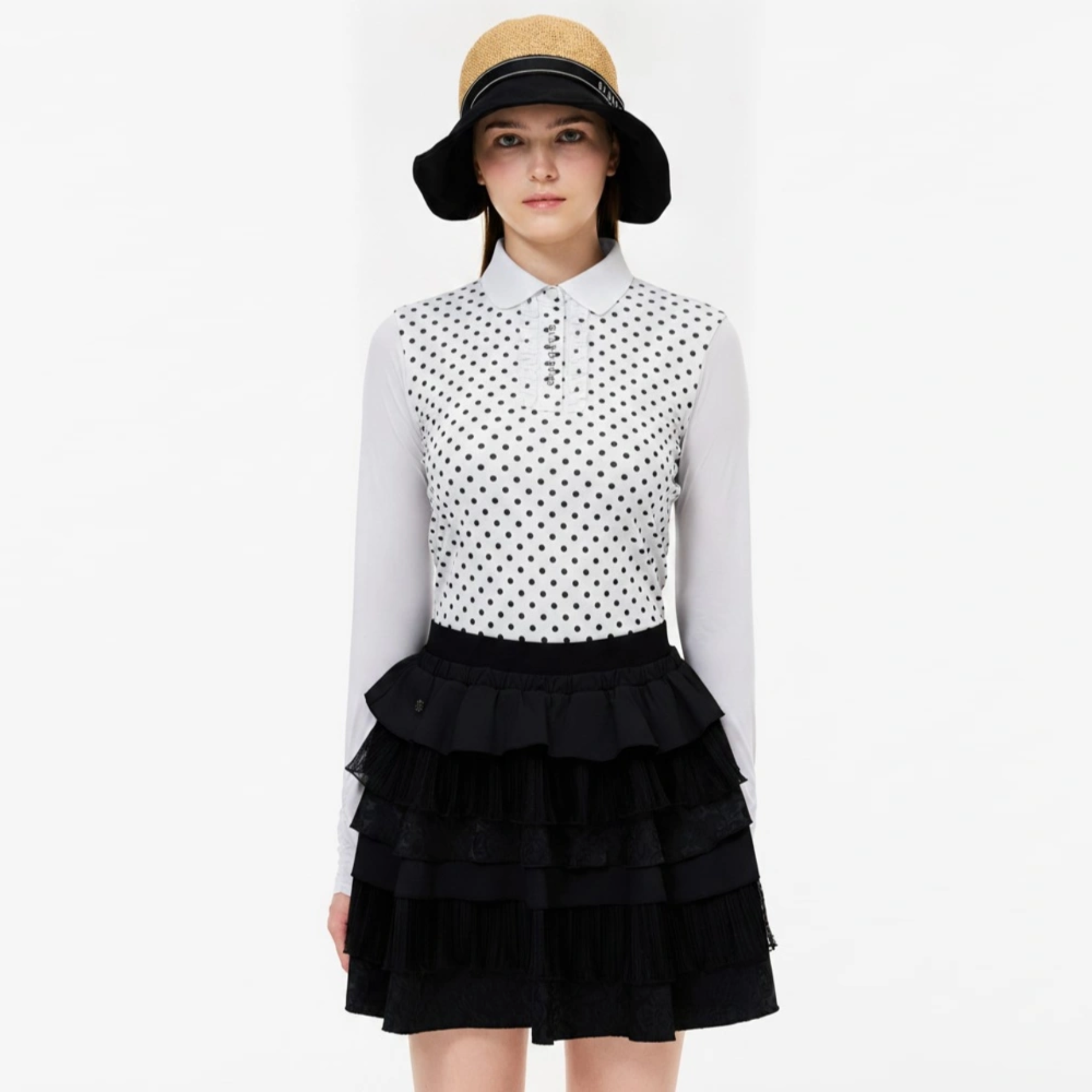 A model wearing a white long-sleeve collared shirt with a black polka dot pattern on the bodice, paired with a black tiered ruffled mini skirt and a tan bucket hat with a black brim.