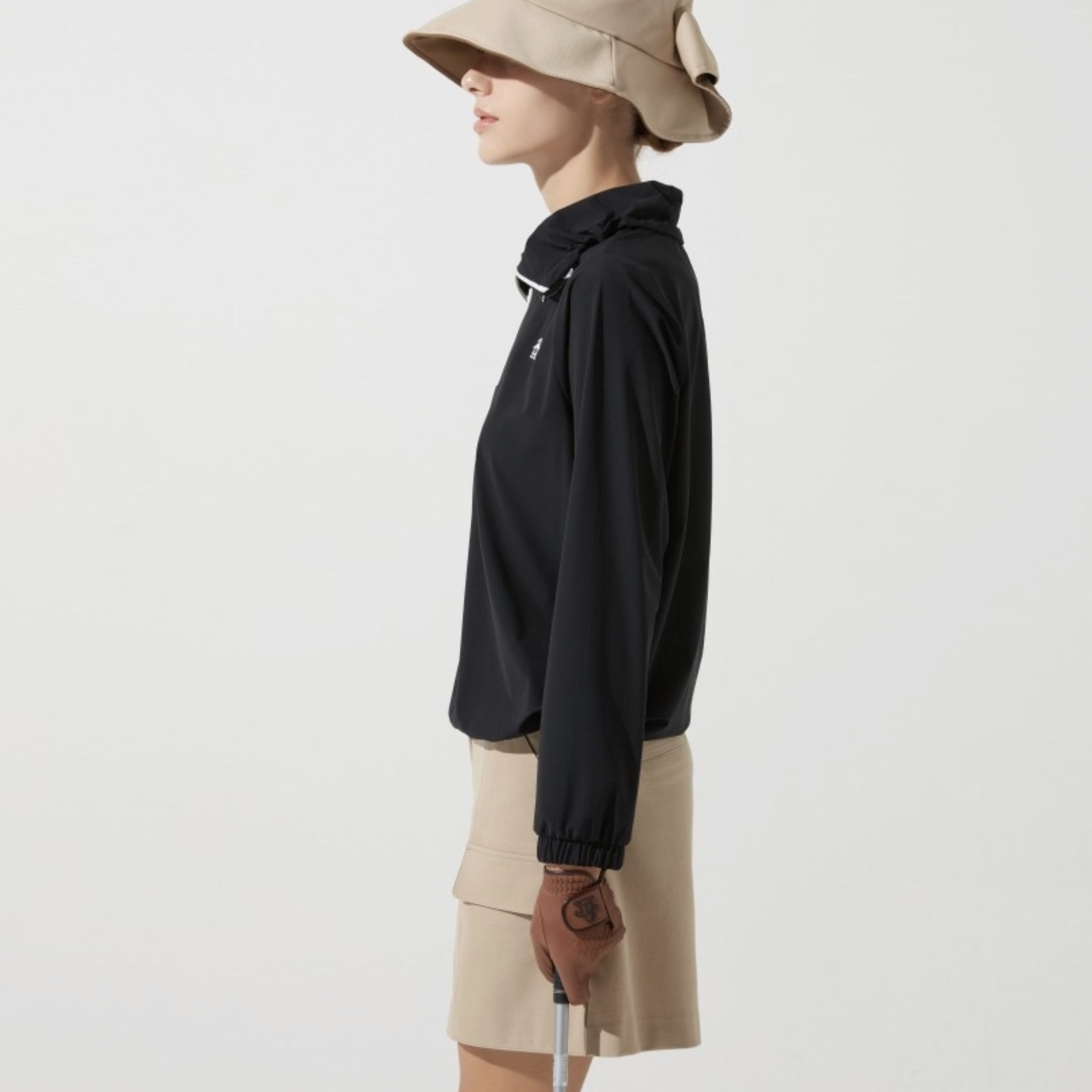 A woman shown in side profile wearing a black lightweight zip-front jacket with a hood and beige skirt, along with a beige wide-brim hat, against a plain light gray studio background.