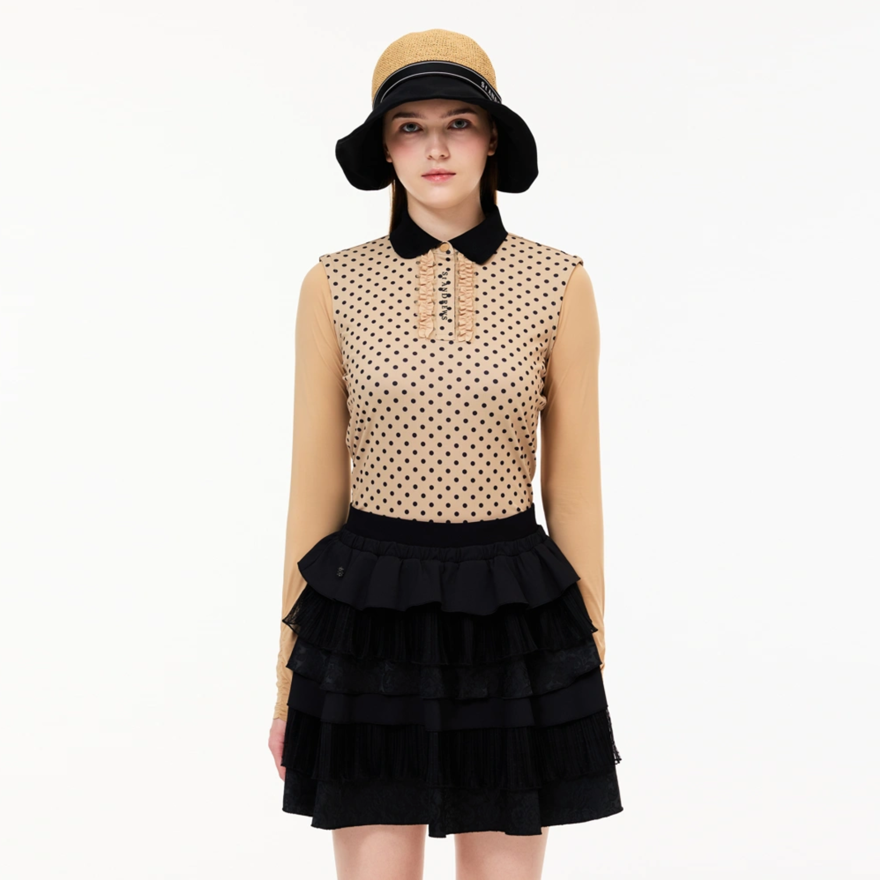 A model wearing a sleeveless beige top with black polka dots and a black collar, paired with a black tiered ruffled skirt and a beige bucket hat with a black band.