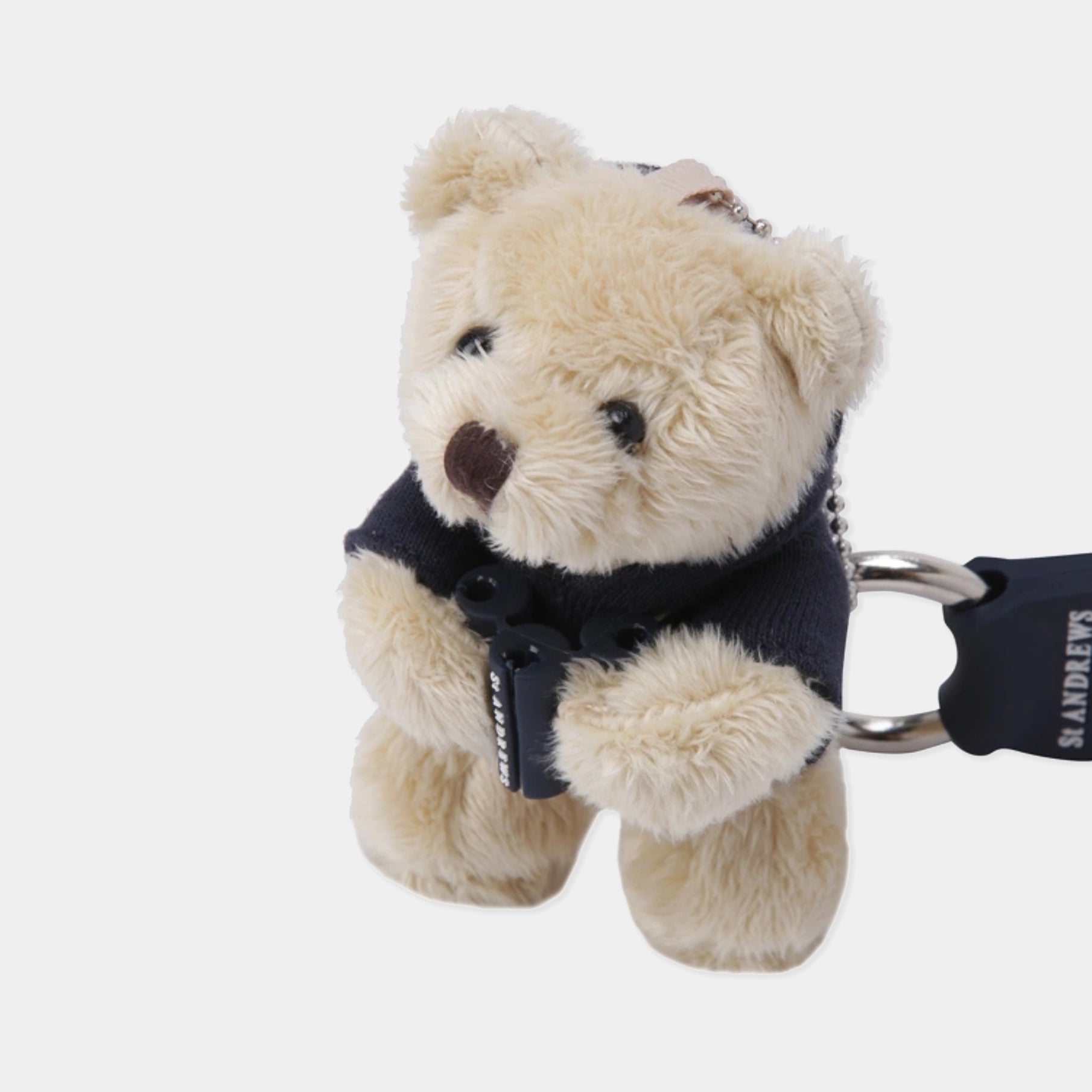 A plush teddy bear keychain in cream-colored faux fur, wearing a navy blue sweater vest and holding a black padlock with a silver ring. The brand name 'ST ANDREWS' is visible on the black strap.