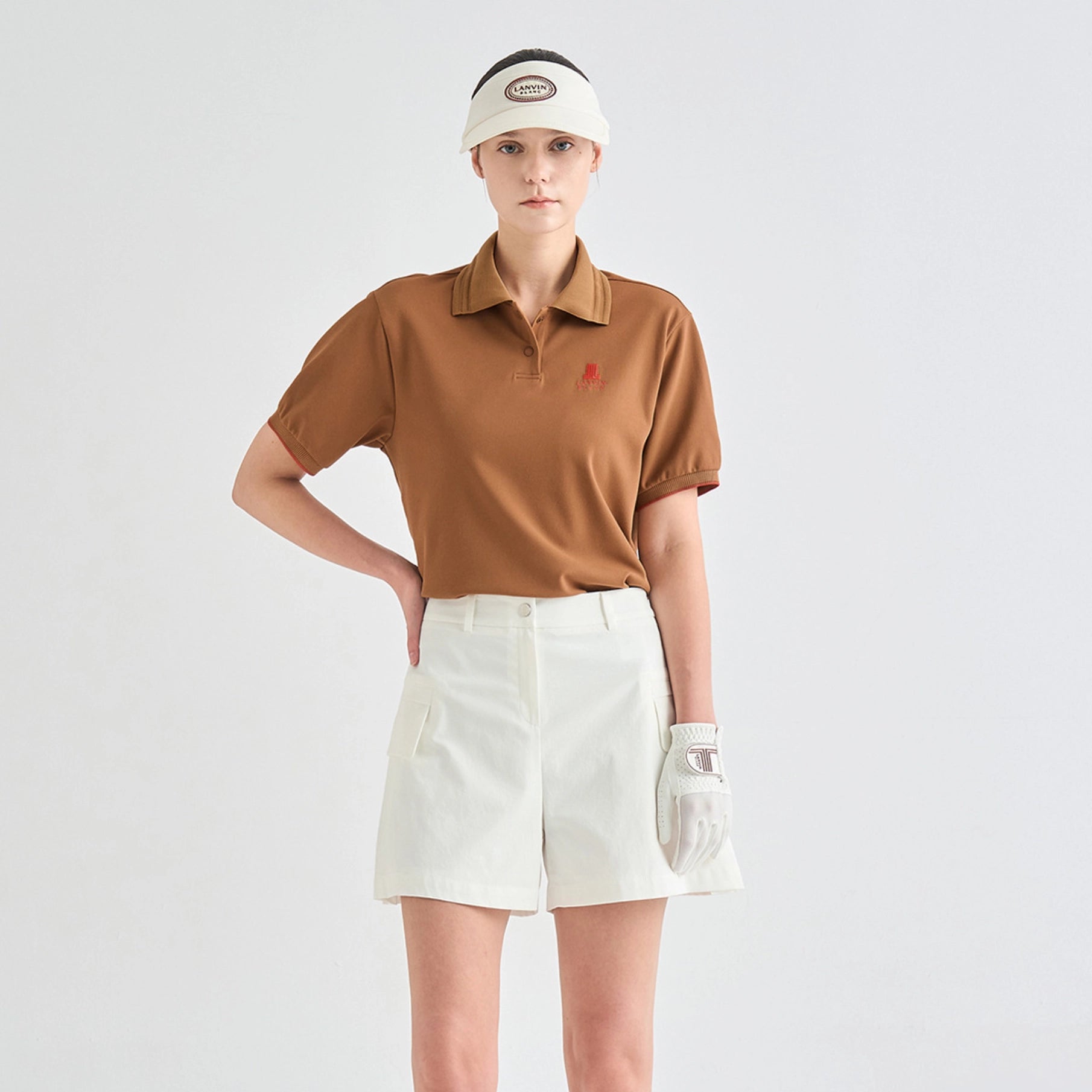 A model wearing a brown short-sleeve polo shirt with a small red embroidered logo on the chest, paired with off-white shorts, a white visor, and a white golf glove, set against a plain light gray studio background.