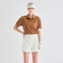 A model wearing a brown short-sleeve polo shirt with a small red embroidered logo on the chest, paired with off-white shorts, a white visor, and a white golf glove, set against a plain light gray studio background.
