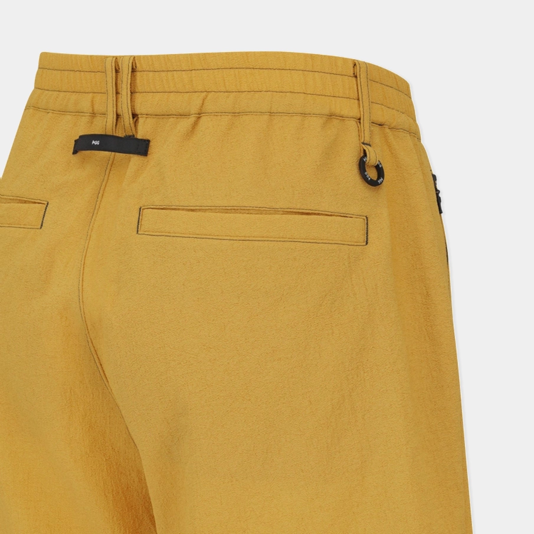 Close-up of the back waistband and rear pockets of mustard yellow trousers, showing a black fabric brand tab and a small black metal ring detail on a light gray background.